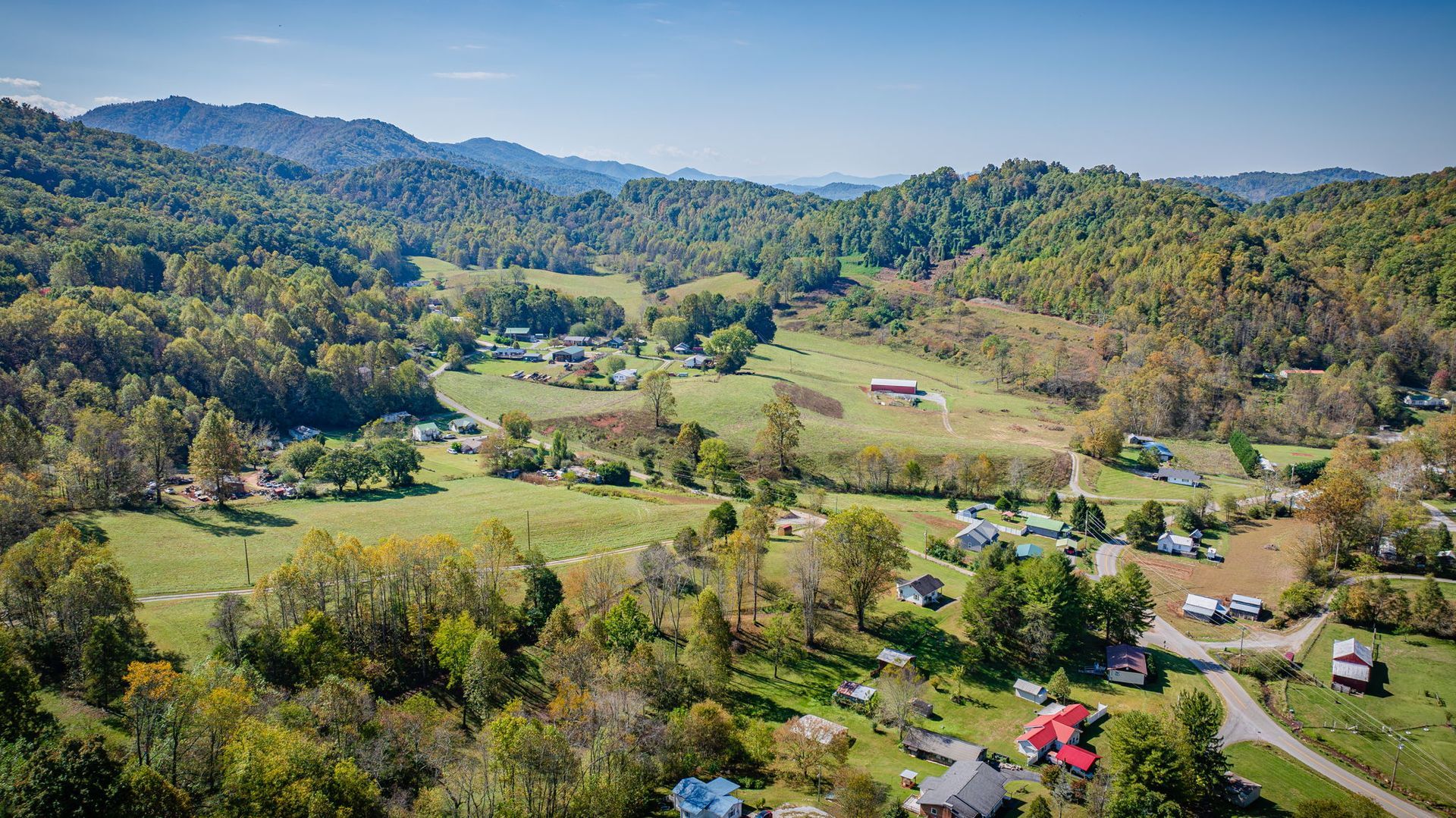 Aerial view of a lush green valley with scattered houses, winding road, and forested hills under clear blue sky