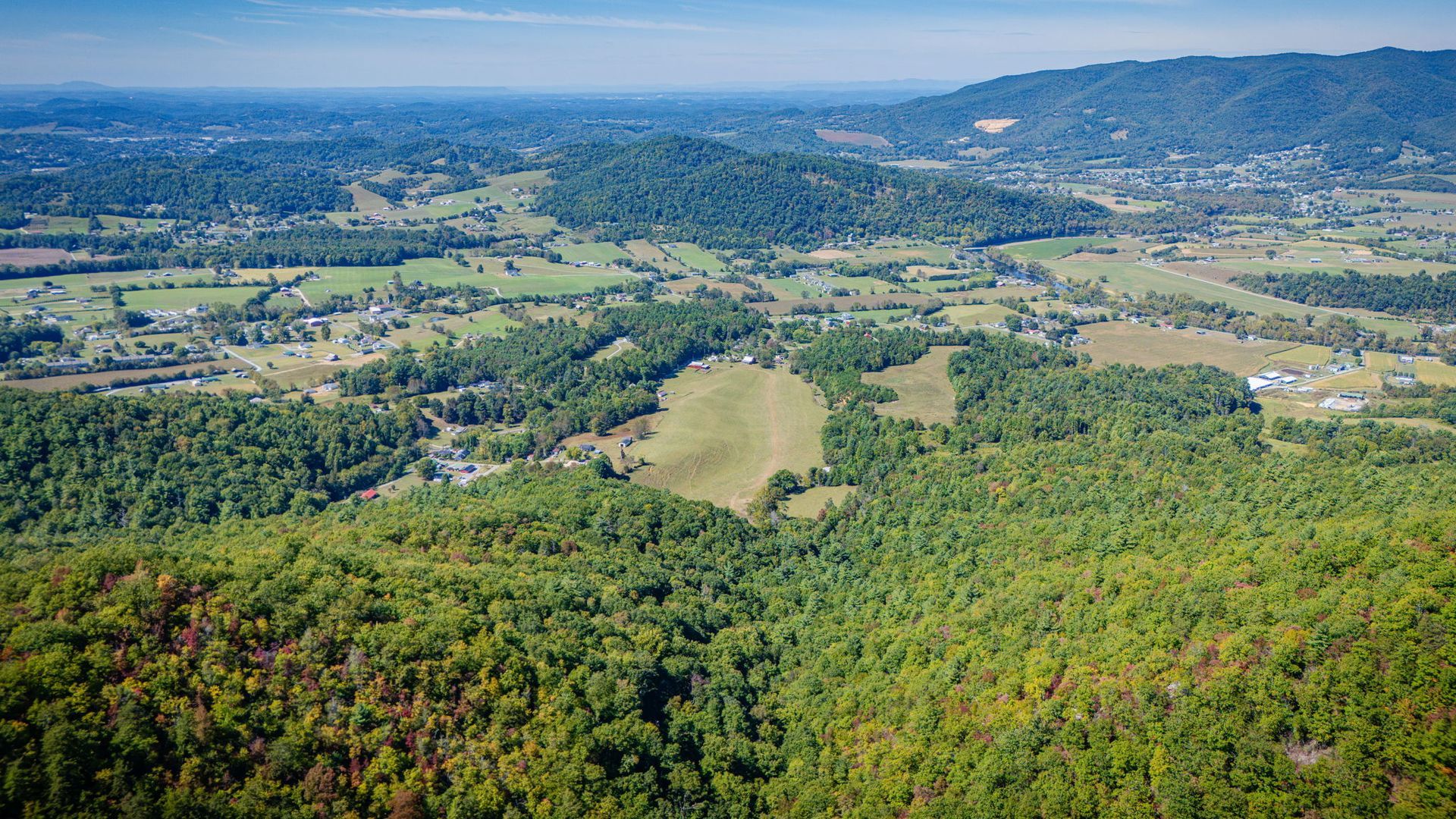 Aerial view of a green valley with fields, forested hills, and distant mountains under a clear blue sky