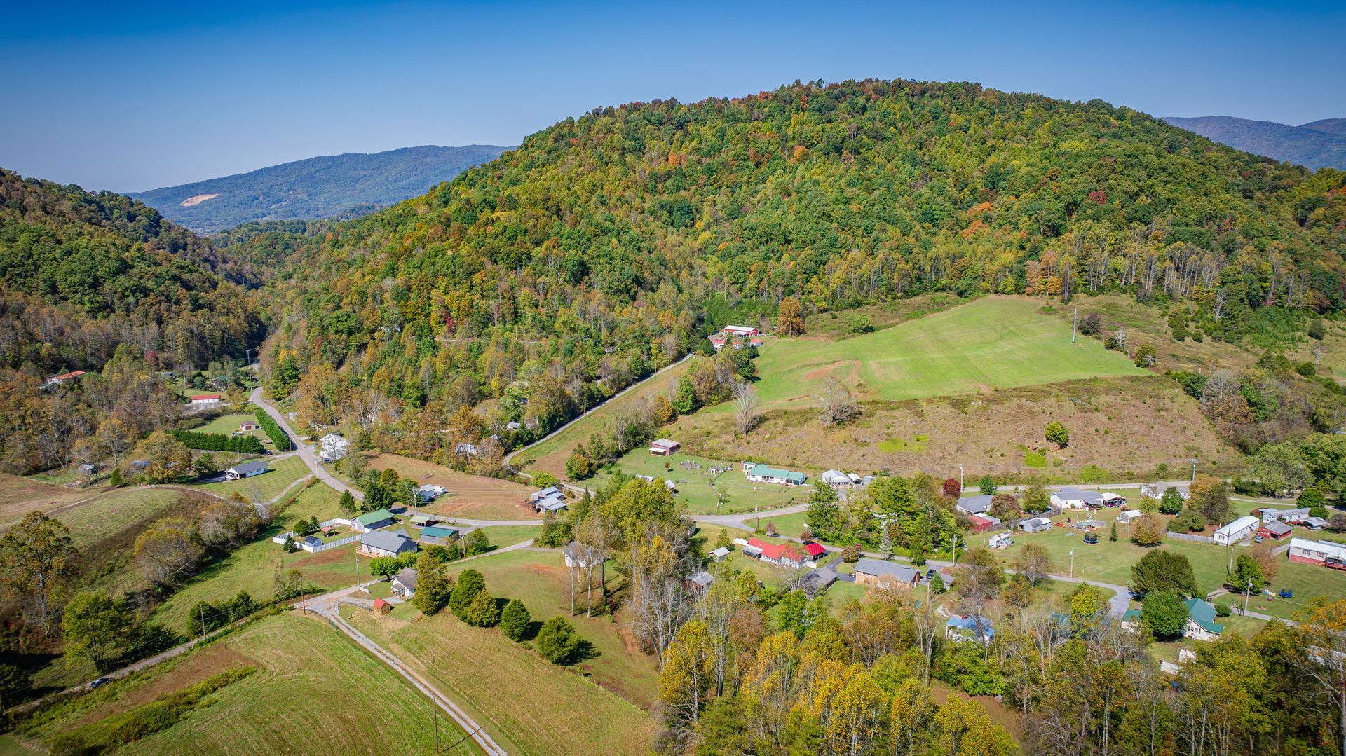 Aerial view of a rural valley with green hills, scattered houses, fields, and a winding road under clear skies.