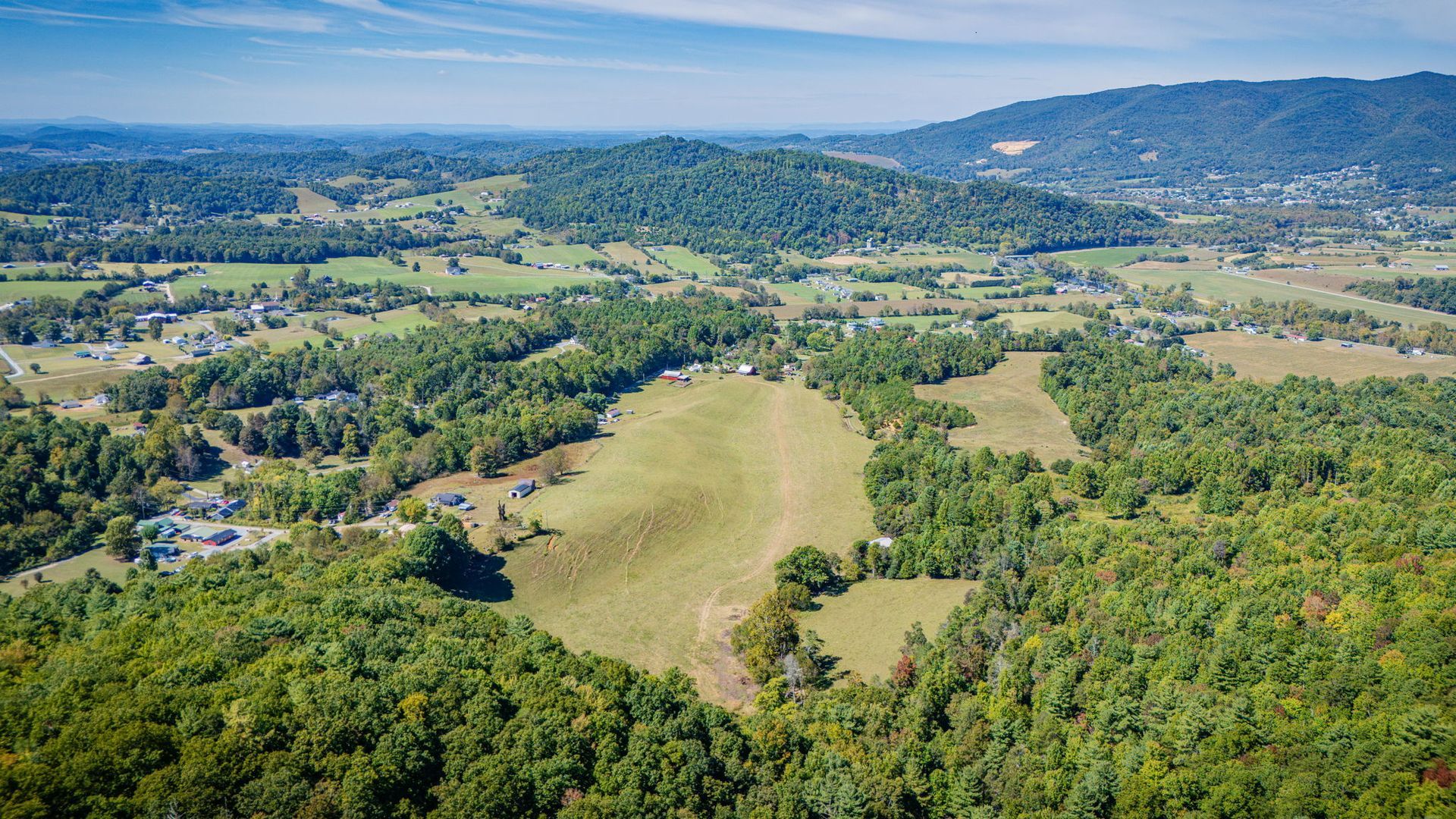 Aerial view of a green valley with open fields, dense trees, and distant hills under a blue sky.