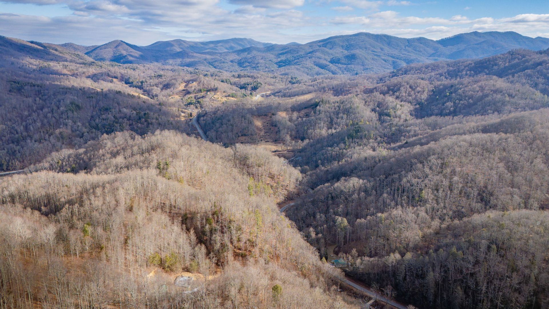 Mountainous landscape with bare trees, valley, and distant hills under a partly cloudy sky.
