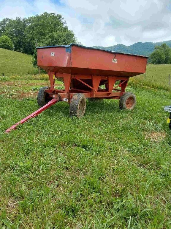 Red farm wagon with one long red hitch parked in a grassy field with trees and hills in the background