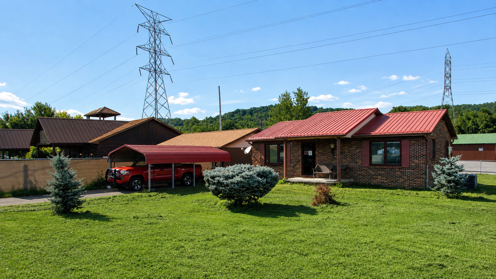 Rural house with red roof, carport, and green lawn under a blue sky