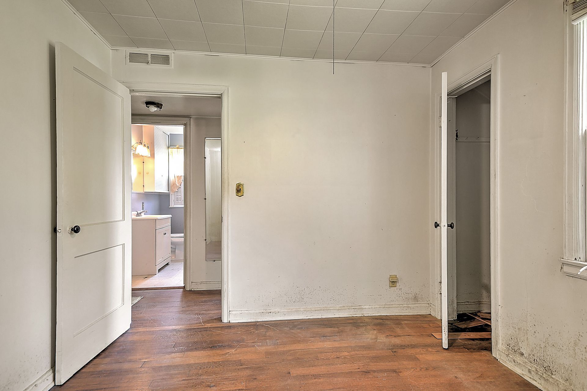 Empty room with white walls, worn wooden floor, and open doors leading to a bright kitchen area