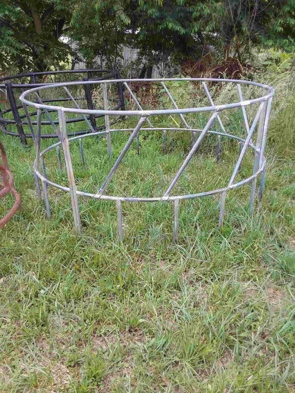Metal livestock feeder frame in a grassy yard, with a rusted red object at the left edge.