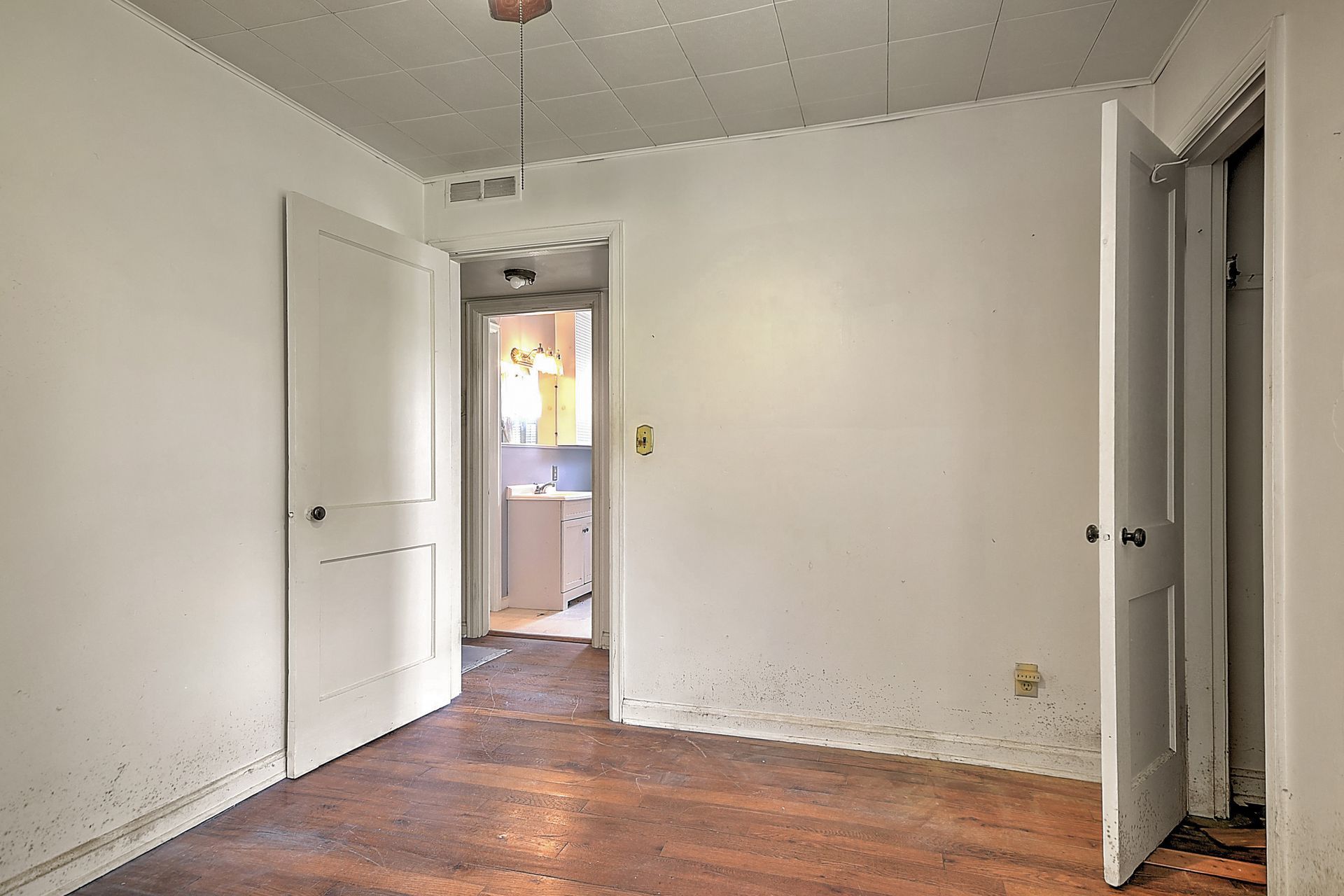 Empty white room with hardwood floor, open doorways, and natural light from an adjoining room.