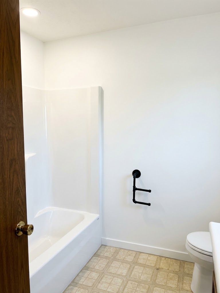 Small white bathroom with tub, toilet, and black toilet paper holder on patterned tile floor.