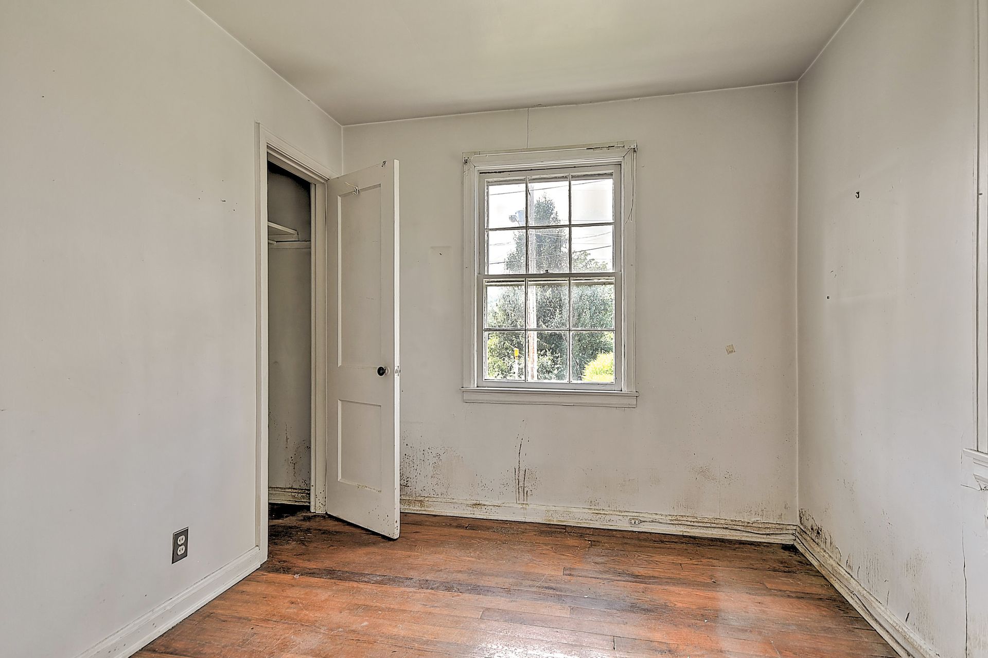 Empty white room with worn wooden floor, open door, and small window letting in daylight