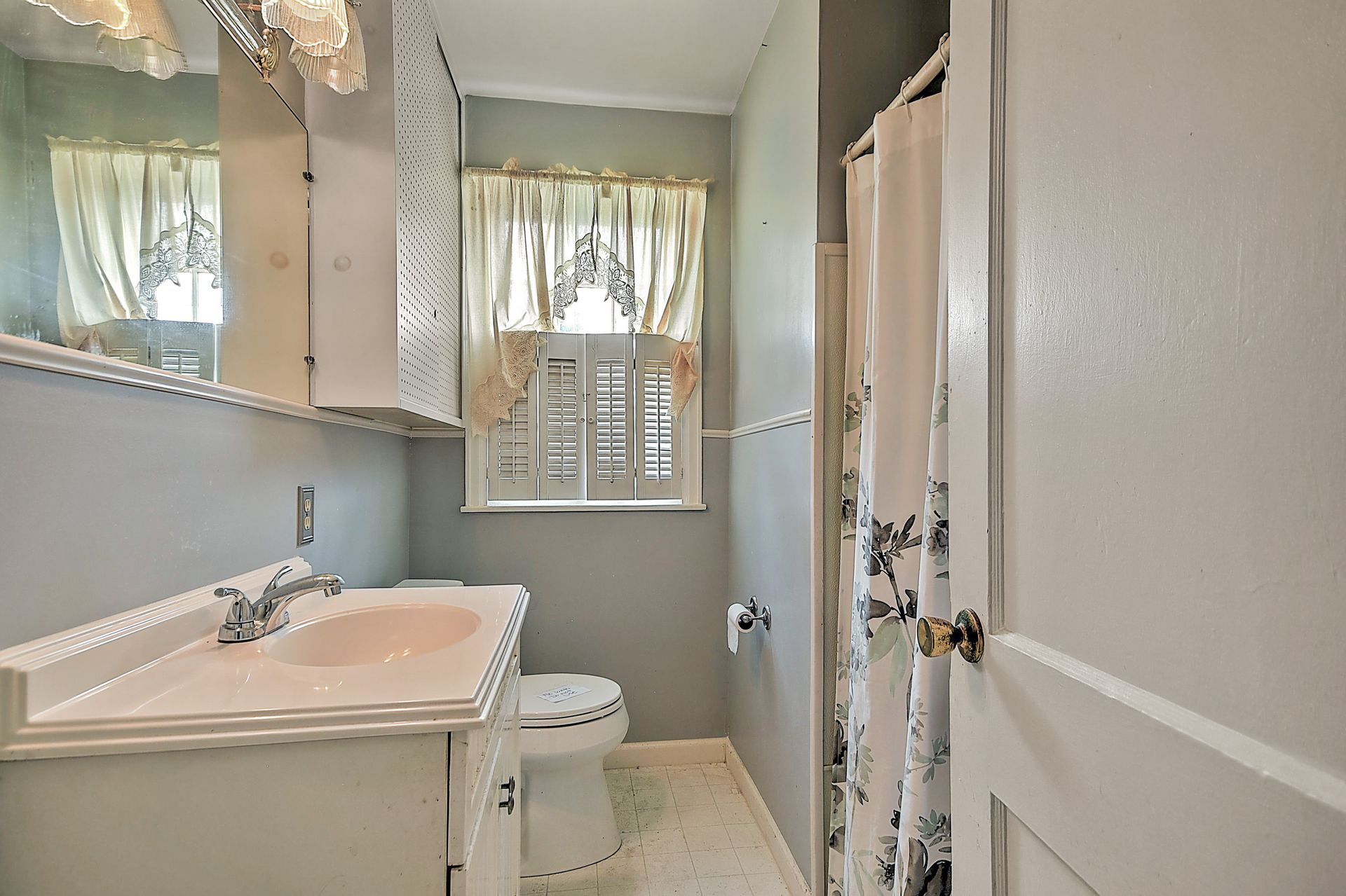 Small bathroom with sink, toilet, mirror cabinet, and a window with sheer curtains.