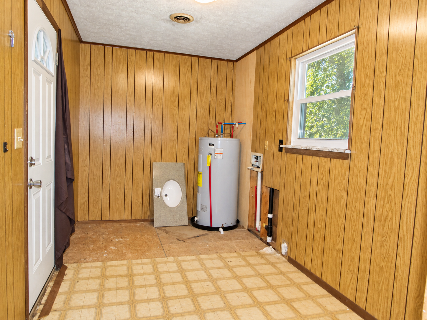 Empty utility room with wood-paneled walls, white door, window, and water heater on a checkered floor.