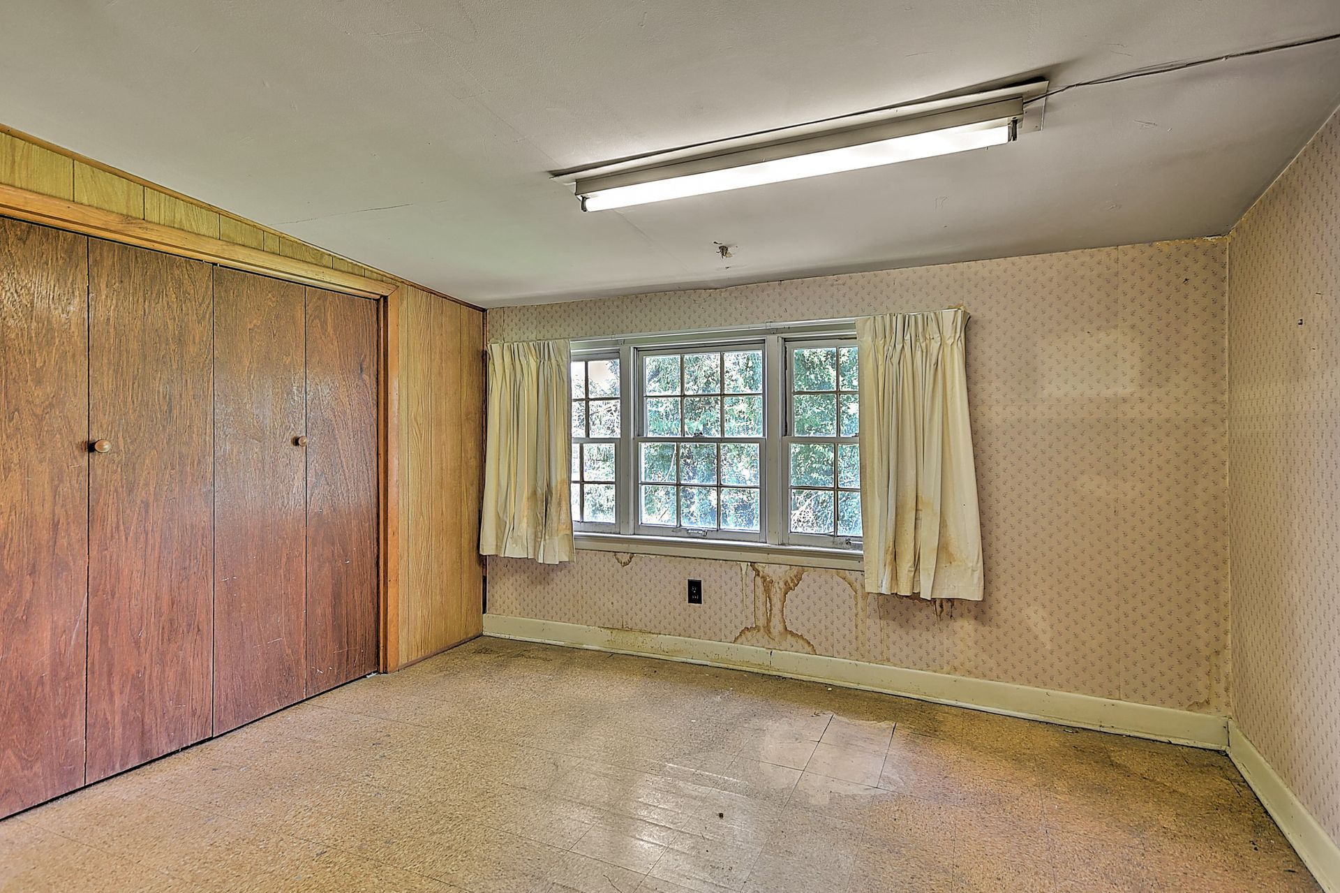 Empty beige room with wood-paneled closet, window with curtains, and fluorescent ceiling light.