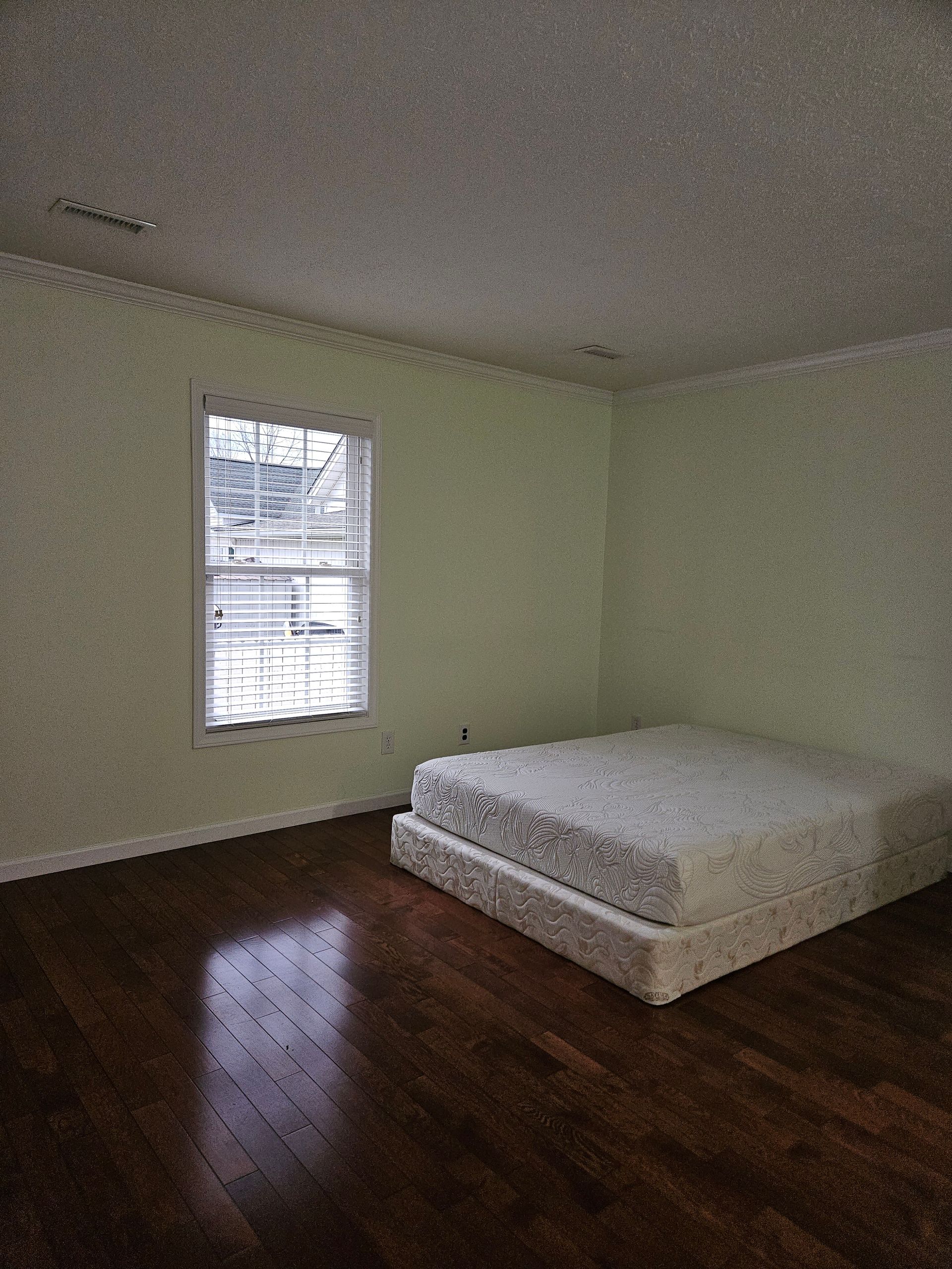 An empty room with pale green walls, dark wood floors, and a mattress on the floor near a single window.