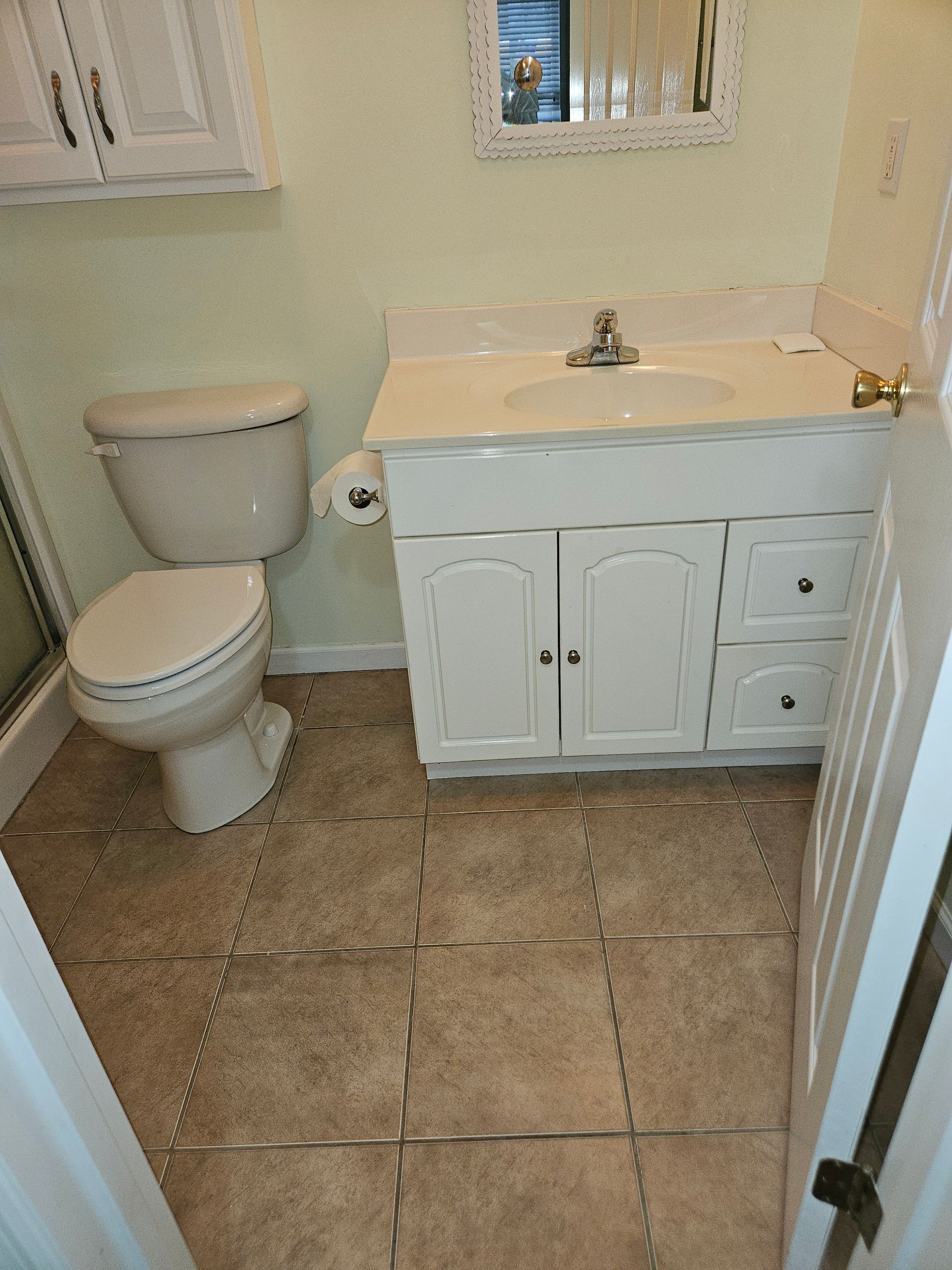 A small bathroom with a white vanity sink, toilet, beige tiled floor, and a mirror against light-colored walls.