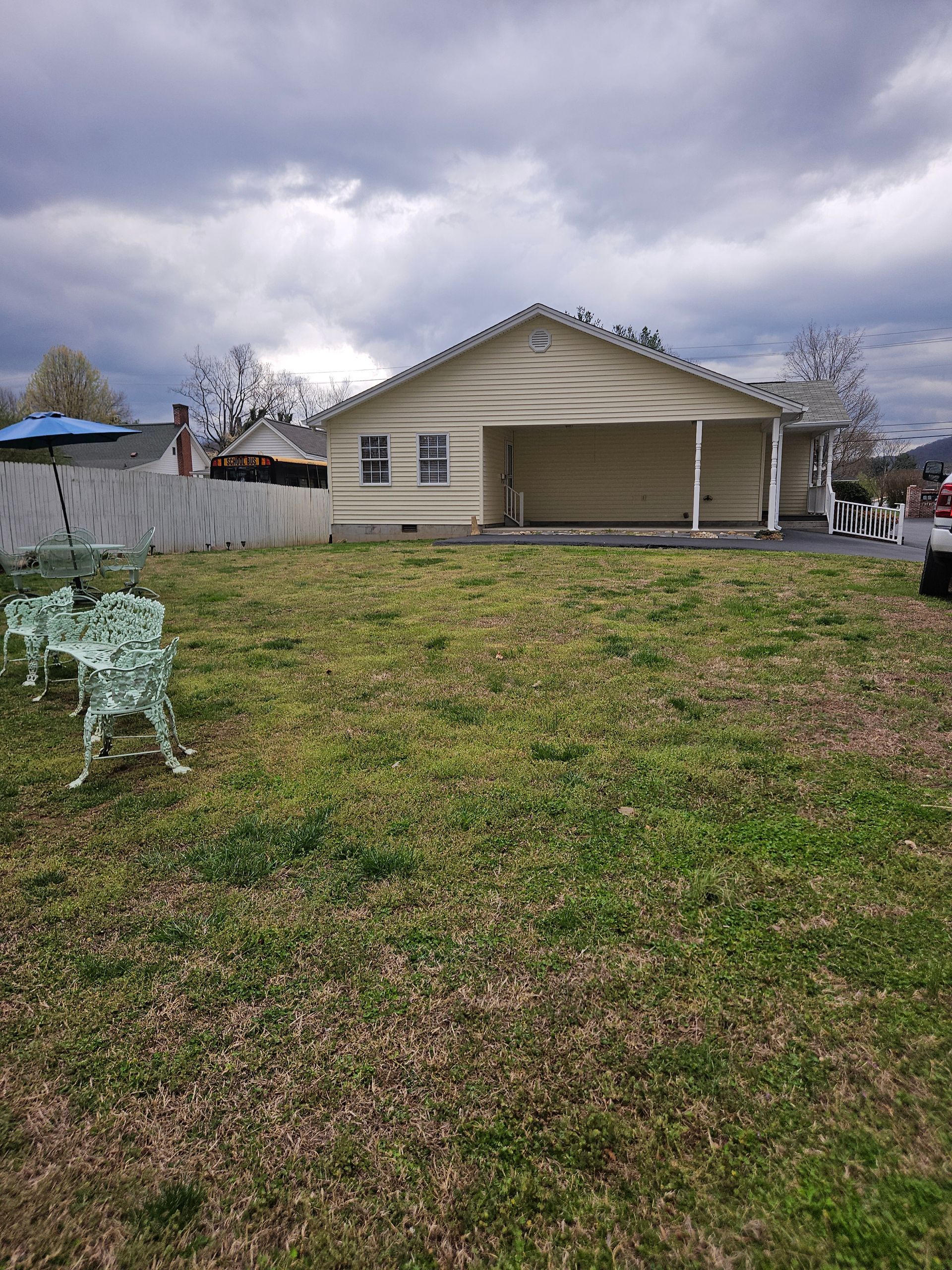 A side view of a beige house with a carport, viewed from a yard with green grass and wrought iron lawn chairs.