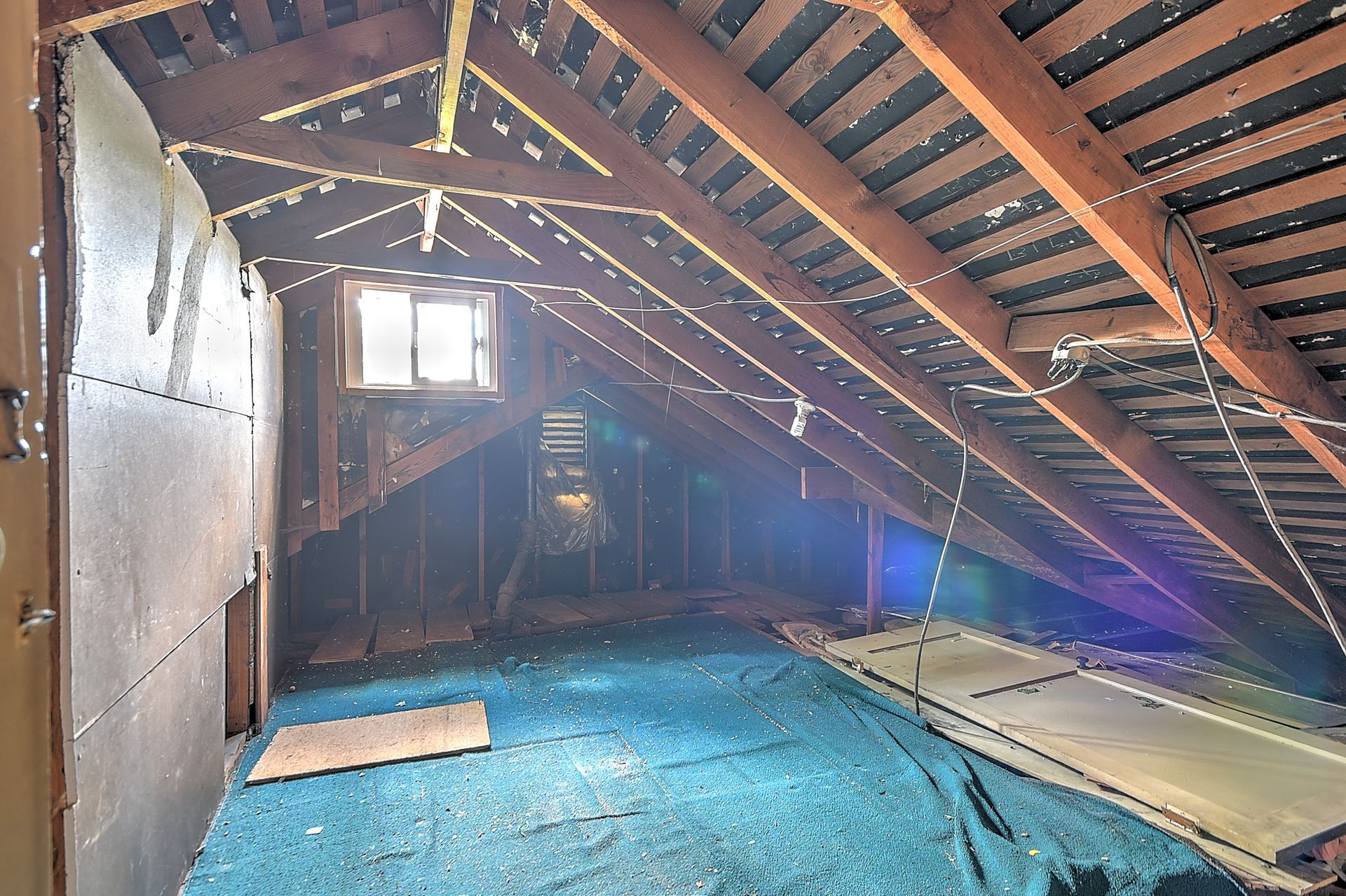Attic interior with exposed rafters, blue carpet, and a small window letting in light.