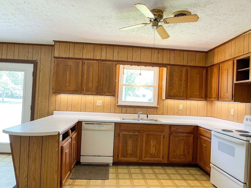 Empty wood-paneled kitchen with white appliances, cabinets, and a ceiling fan