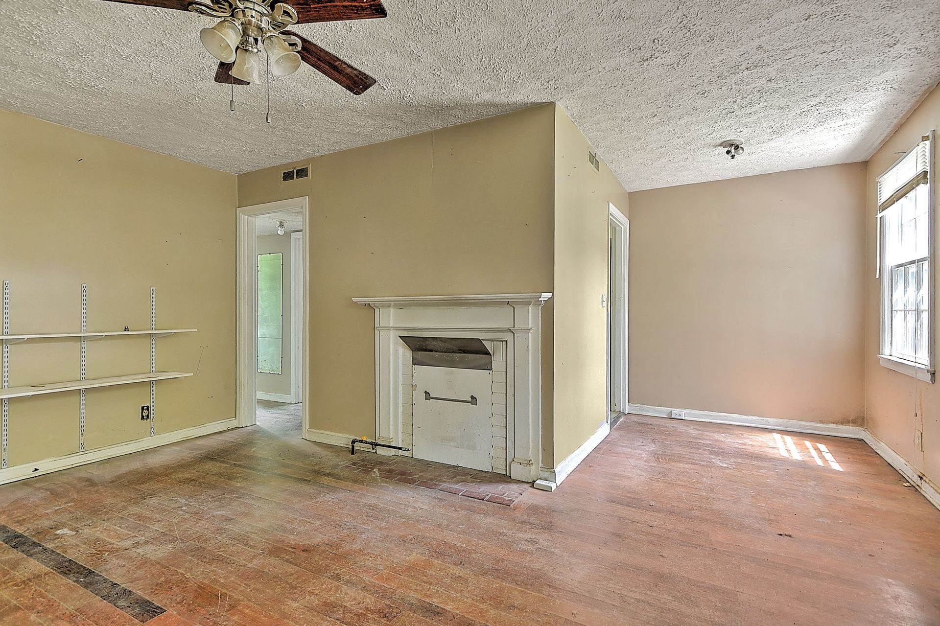 Empty living room with beige walls, fireplace, ceiling fan, and worn carpet near a bright window.