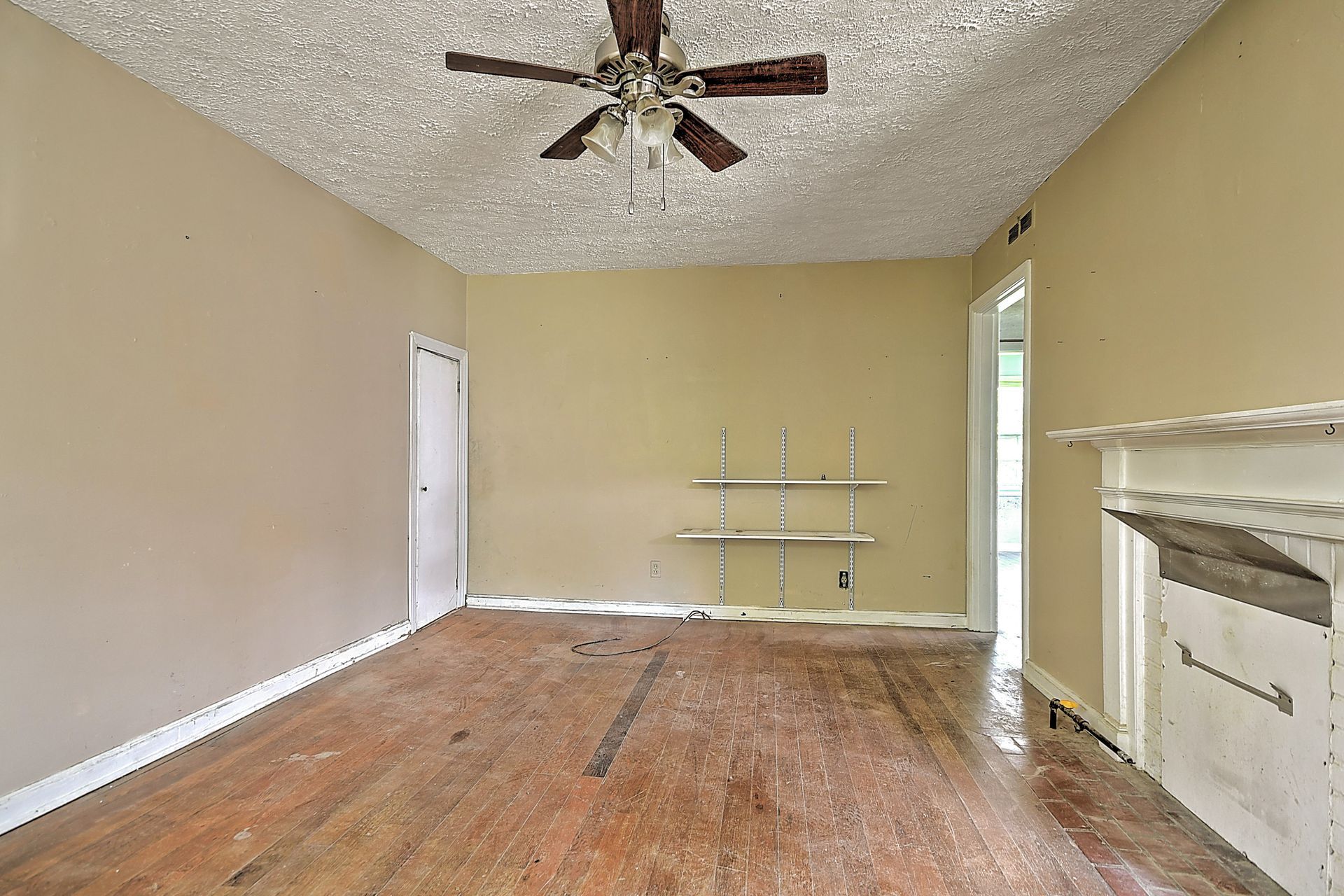 Empty beige living room with hardwood floors, ceiling fan, shelves, and a fireplace mantel