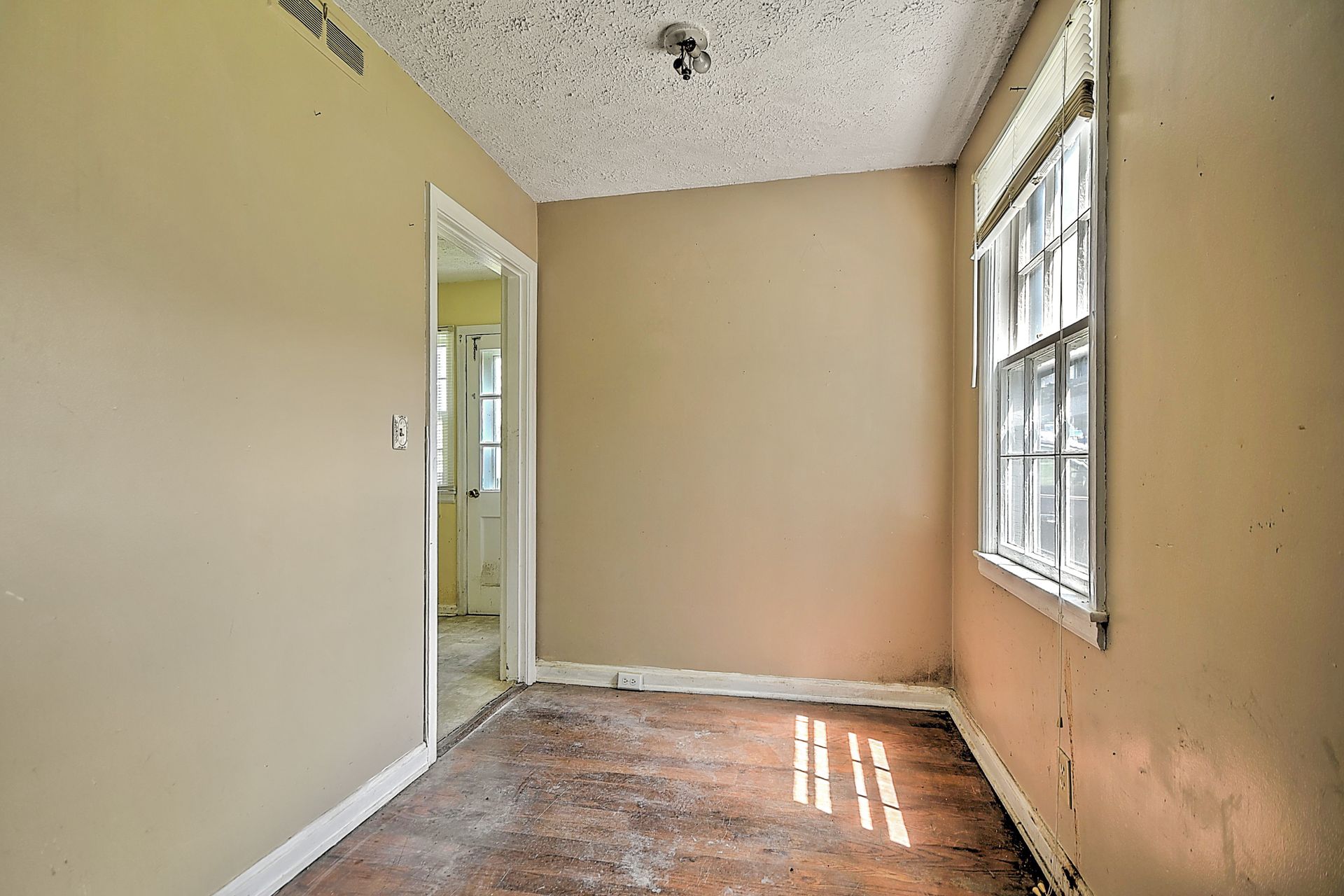 Empty beige room with white trim, worn wood floor, and a window letting in sunlight