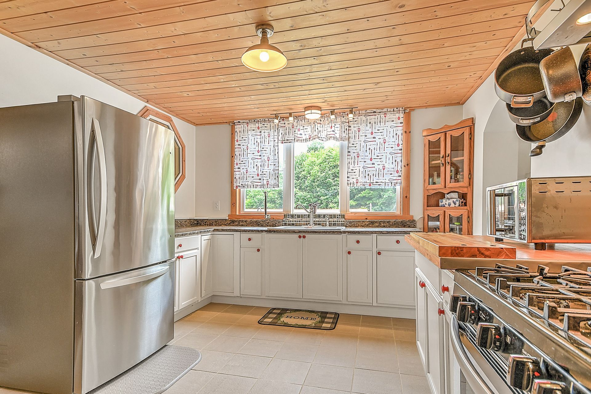Bright kitchen with stainless fridge, white cabinets, wood ceiling, and large window over the sink