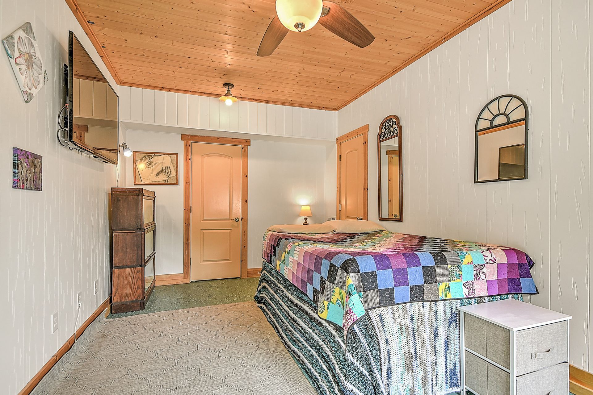 Bedroom with a quilted bedspread, wood ceiling, ceiling fan, and mirrors on white paneled walls.