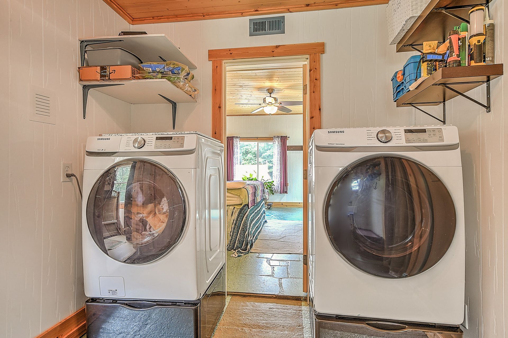 Laundry room with two white front-loading washers, shelves above, and a doorway to a bright room beyond.