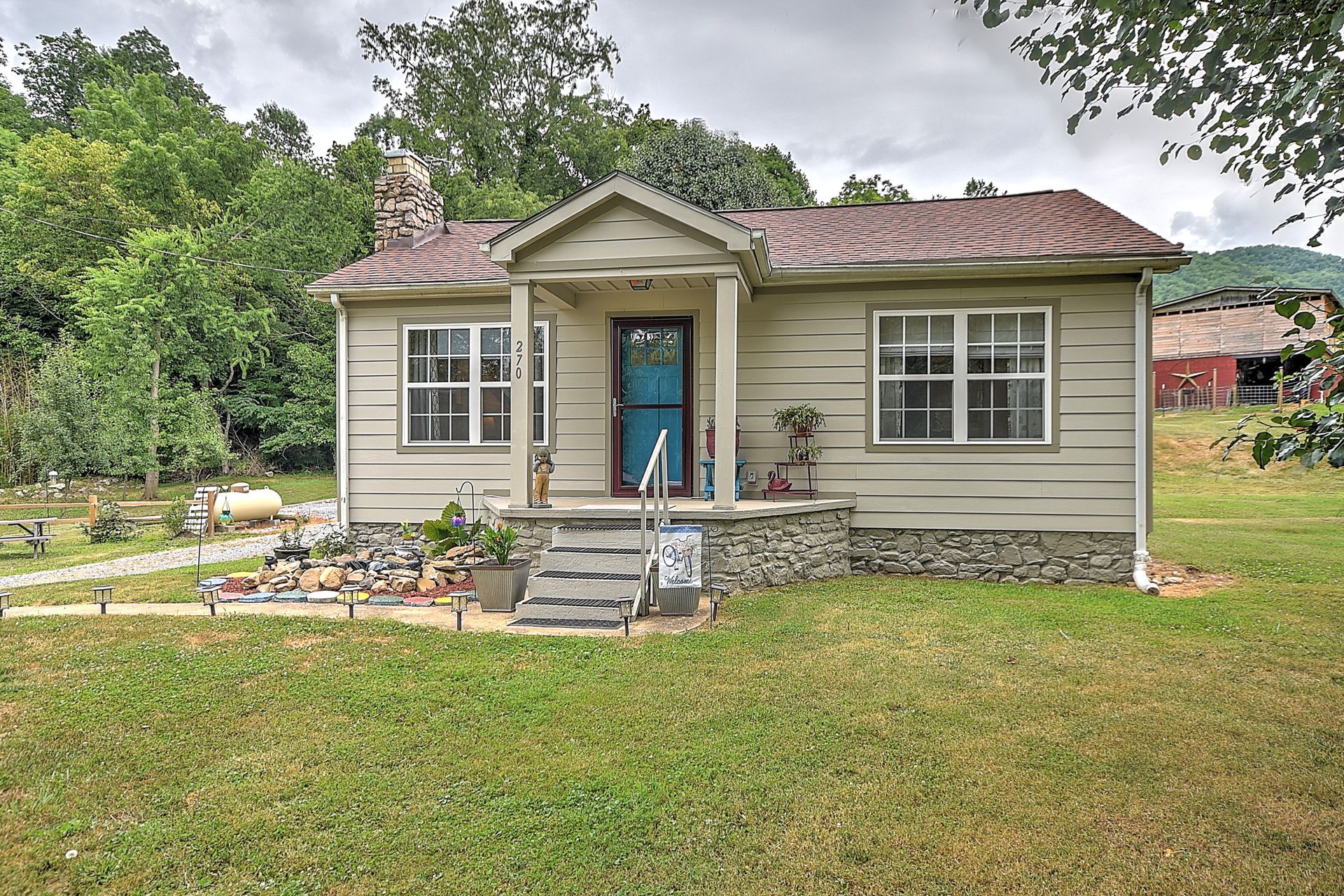 Small beige house with a red roof, blue front door, and flower bed in a grassy yard