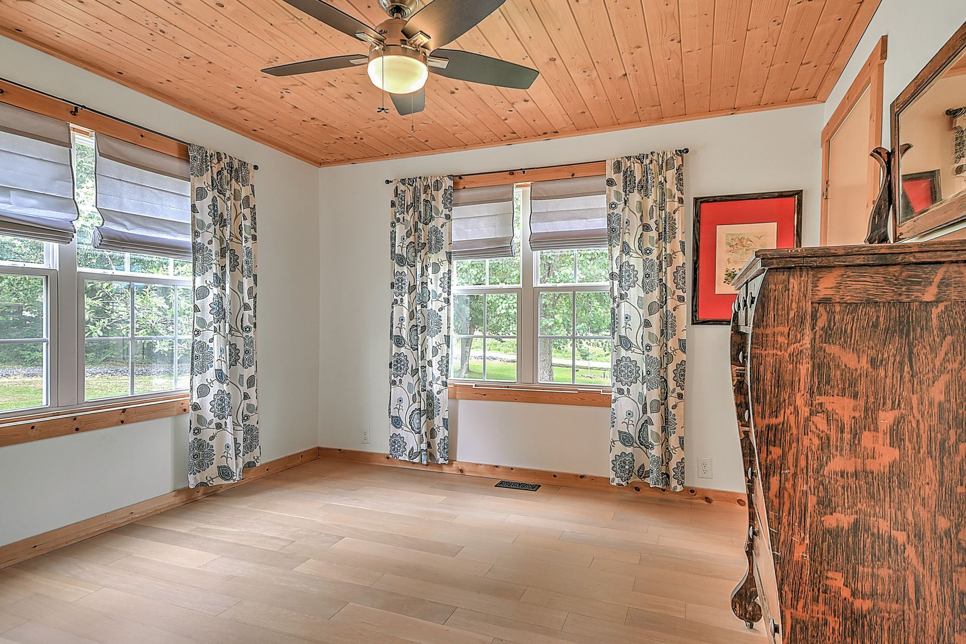 Empty sunlit room with wood ceiling, ceiling fan, large windows, and patterned curtains