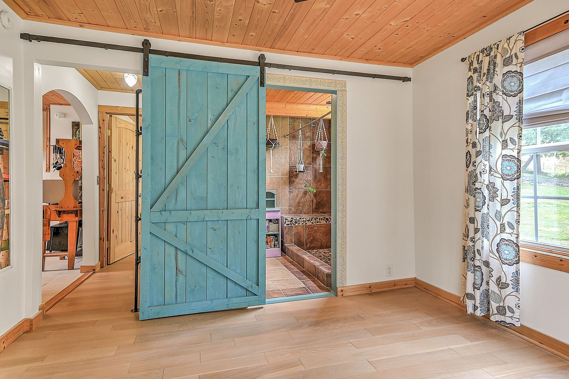 Rustic room with blue sliding barn door, wood ceiling, light walls, and window with patterned curtains