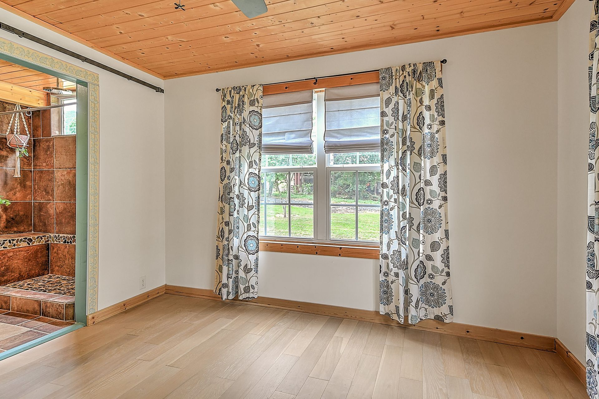Empty room with light wood floors, floral curtains, a window, and an open glass door to outside.