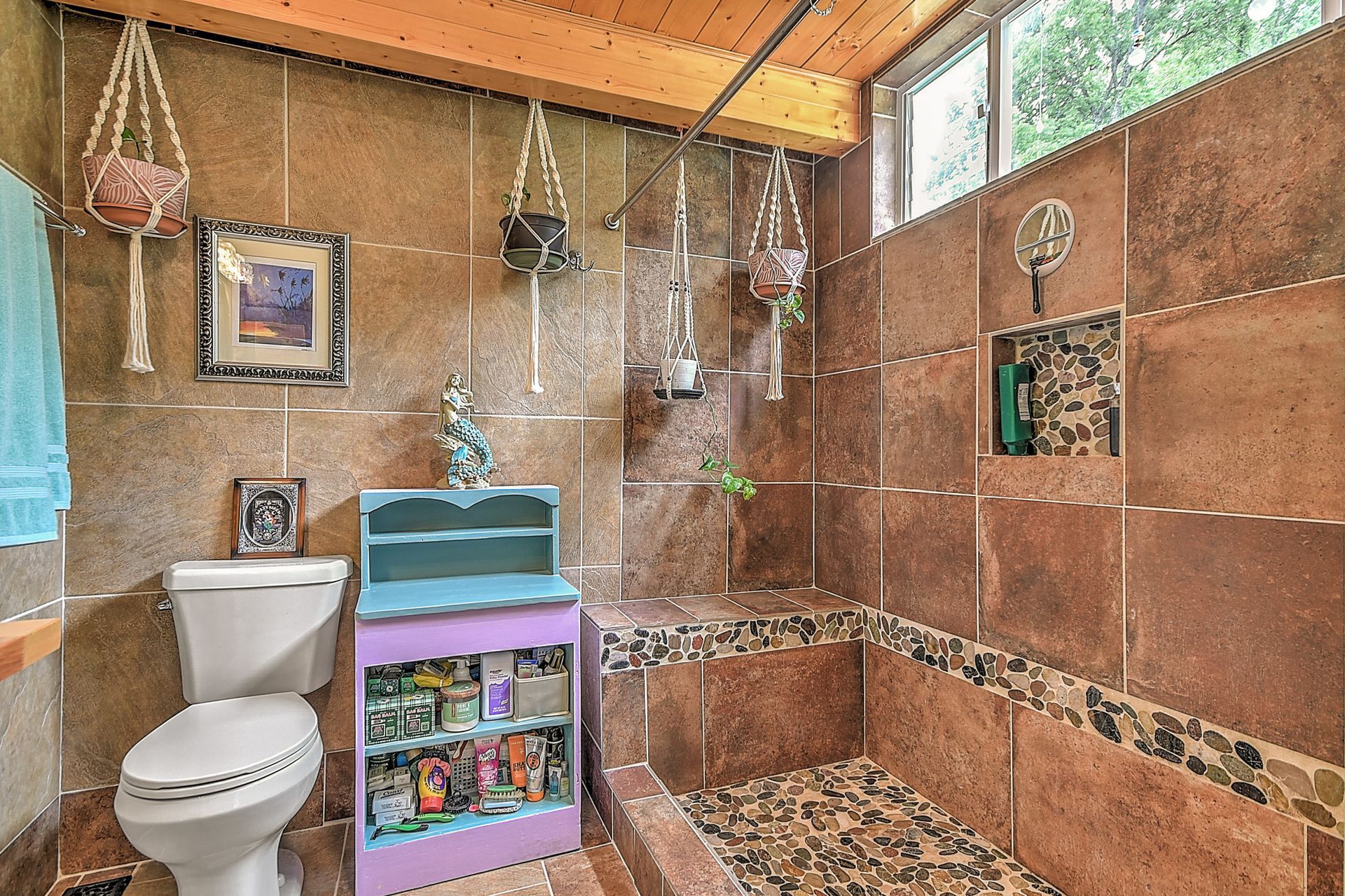 Small rustic bathroom with brown tile walls, white toilet, sink, and a colorful shelf under hanging decor.