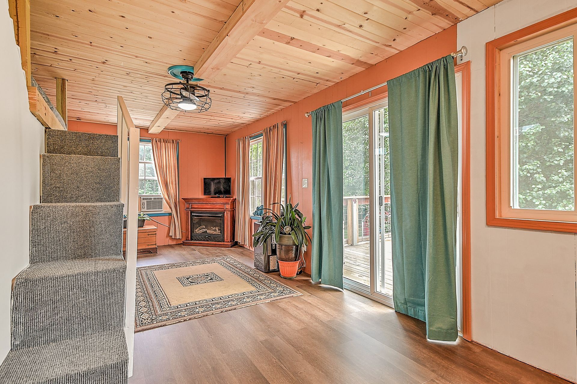 Cozy wood-paneled room with staircase, desk, green curtains, and sliding glass doors overlooking trees