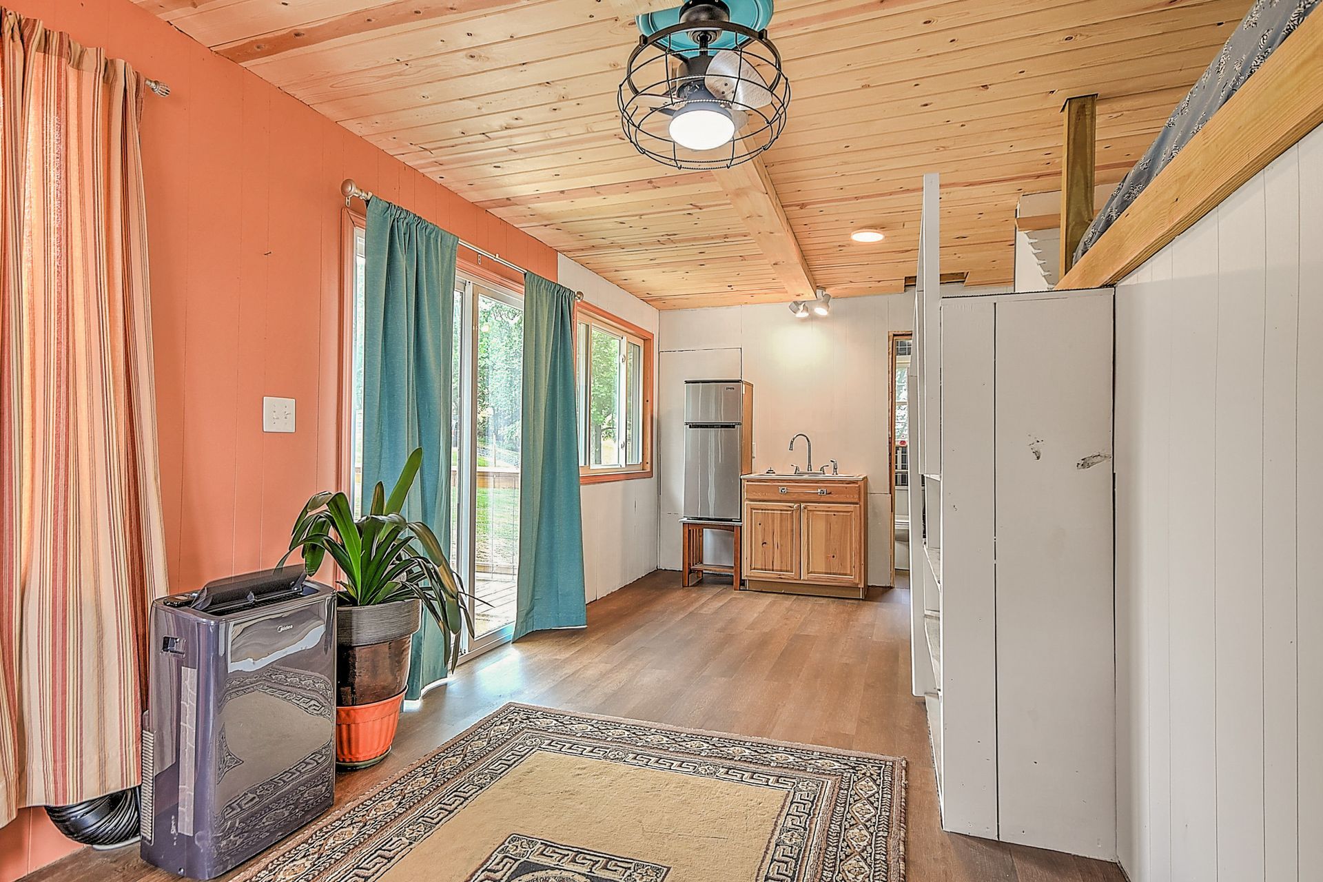 Long, narrow room with wood ceiling, colorful curtains, plants, rug, cabinets, and a stainless-steel fridge