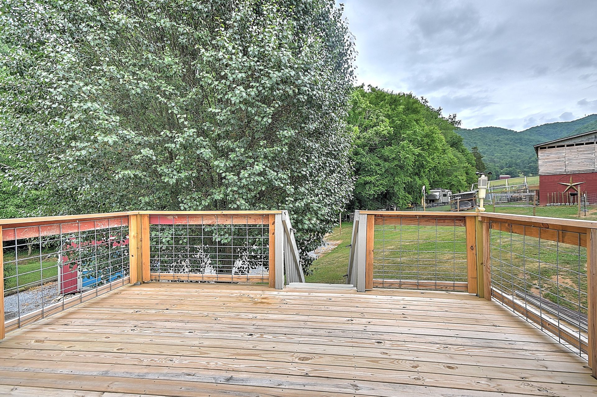 Wooden deck overlooking a fenced yard, large tree, and distant hills under a cloudy sky