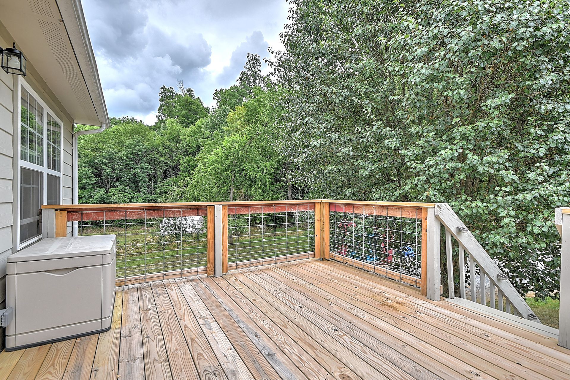 Wooden deck with white railing, gray storage box, and lush green trees beyond.