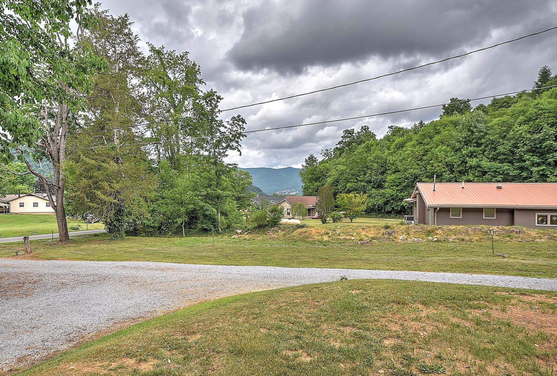 Gravel road beside grassy field and trees, with small house and mountains under cloudy sky.