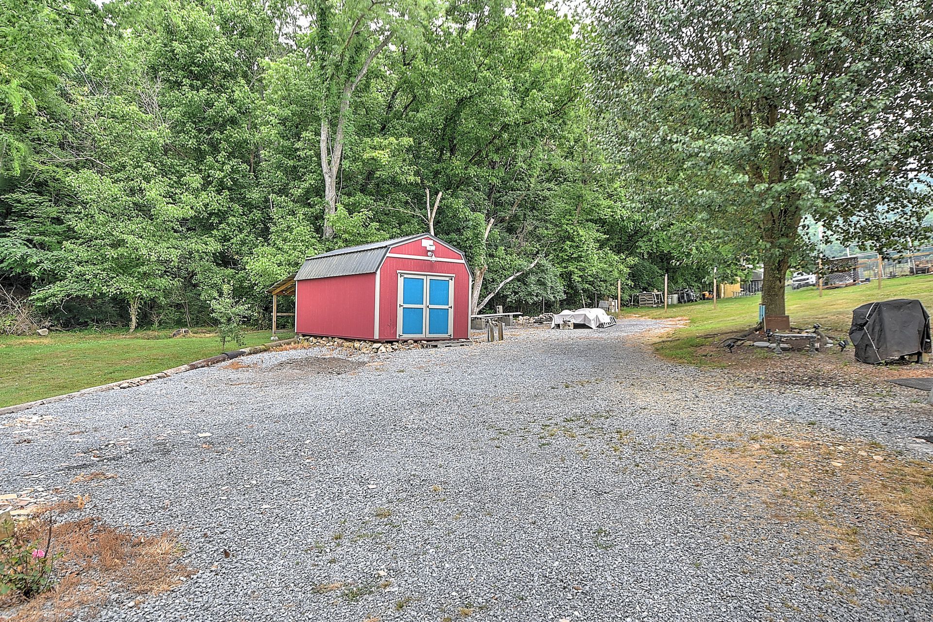 Red shed with blue doors in a gravel driveway, surrounded by green trees and lawn.