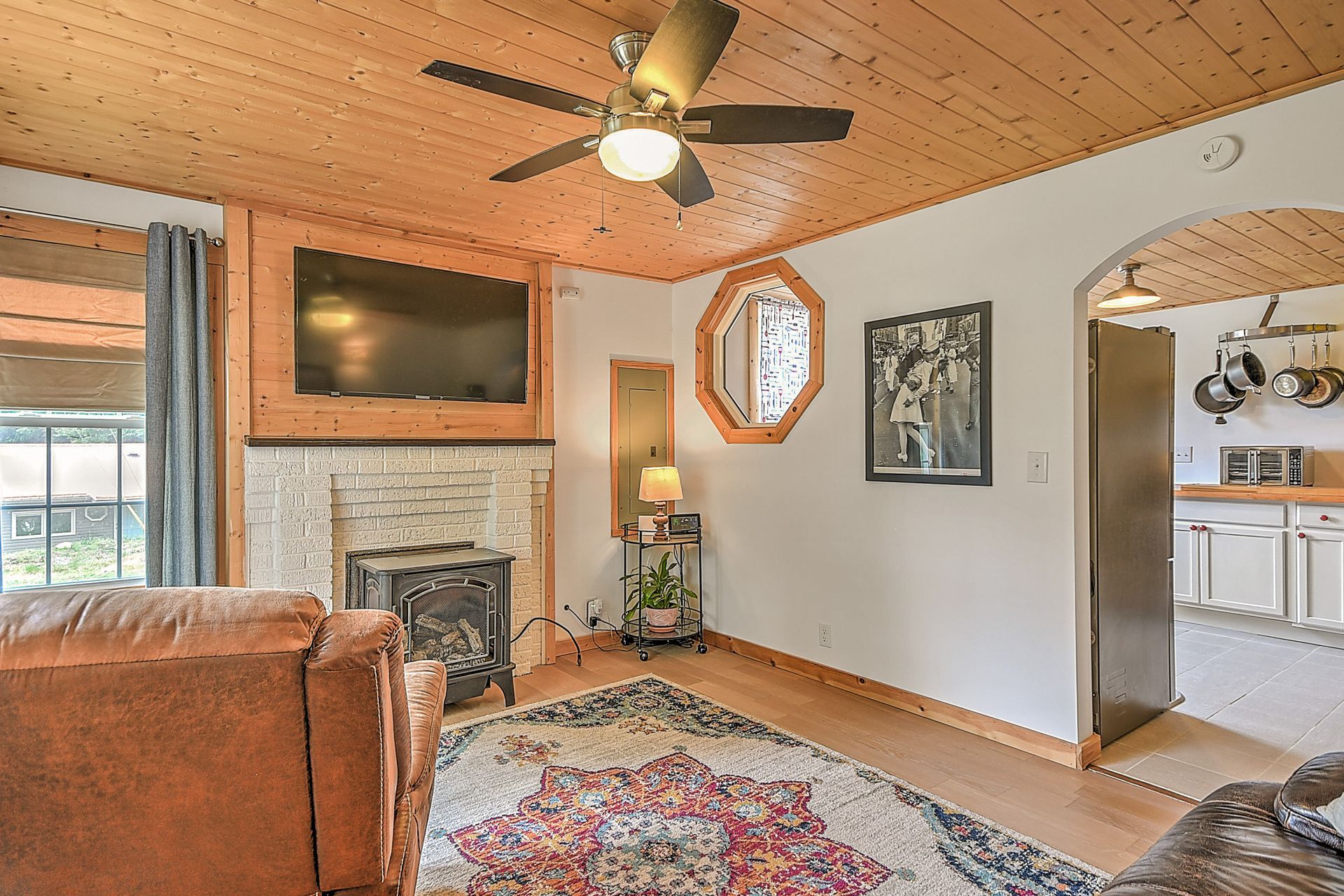 Cozy living room with wood ceiling, fireplace, wall TV, patterned rug, and view into a bright kitchen