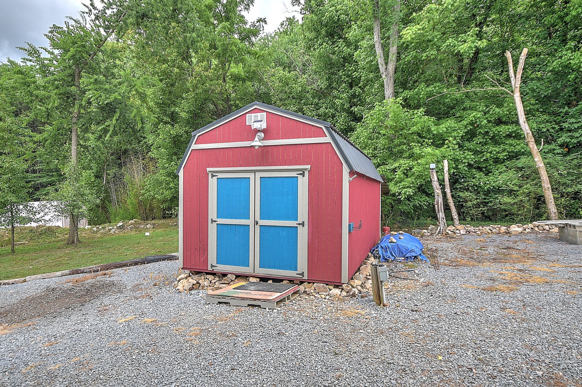 Red garden shed with blue double doors on a gravel lot, surrounded by green trees.