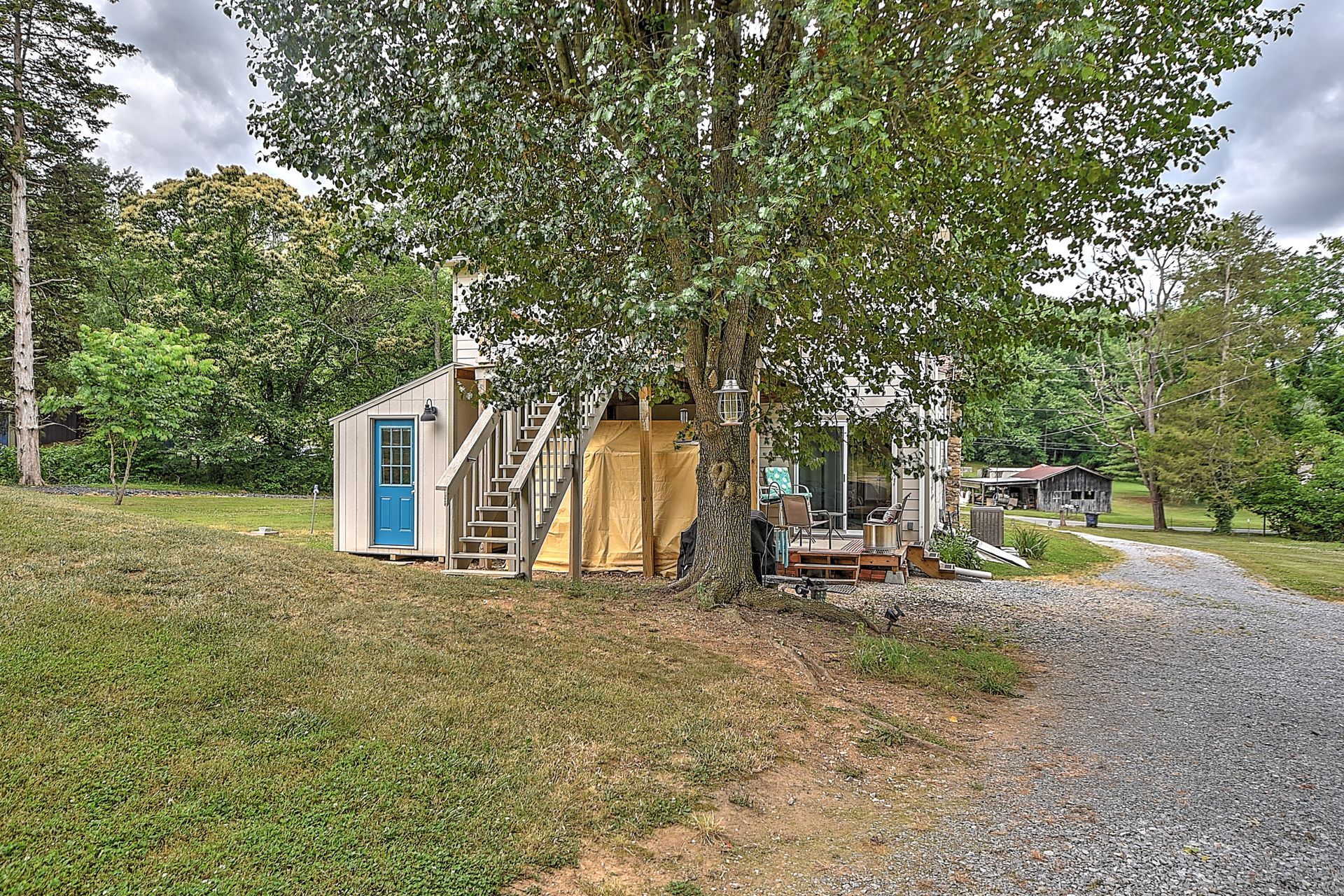 Small blue-and-tan shed under trees beside a gravel driveway in a rural yard