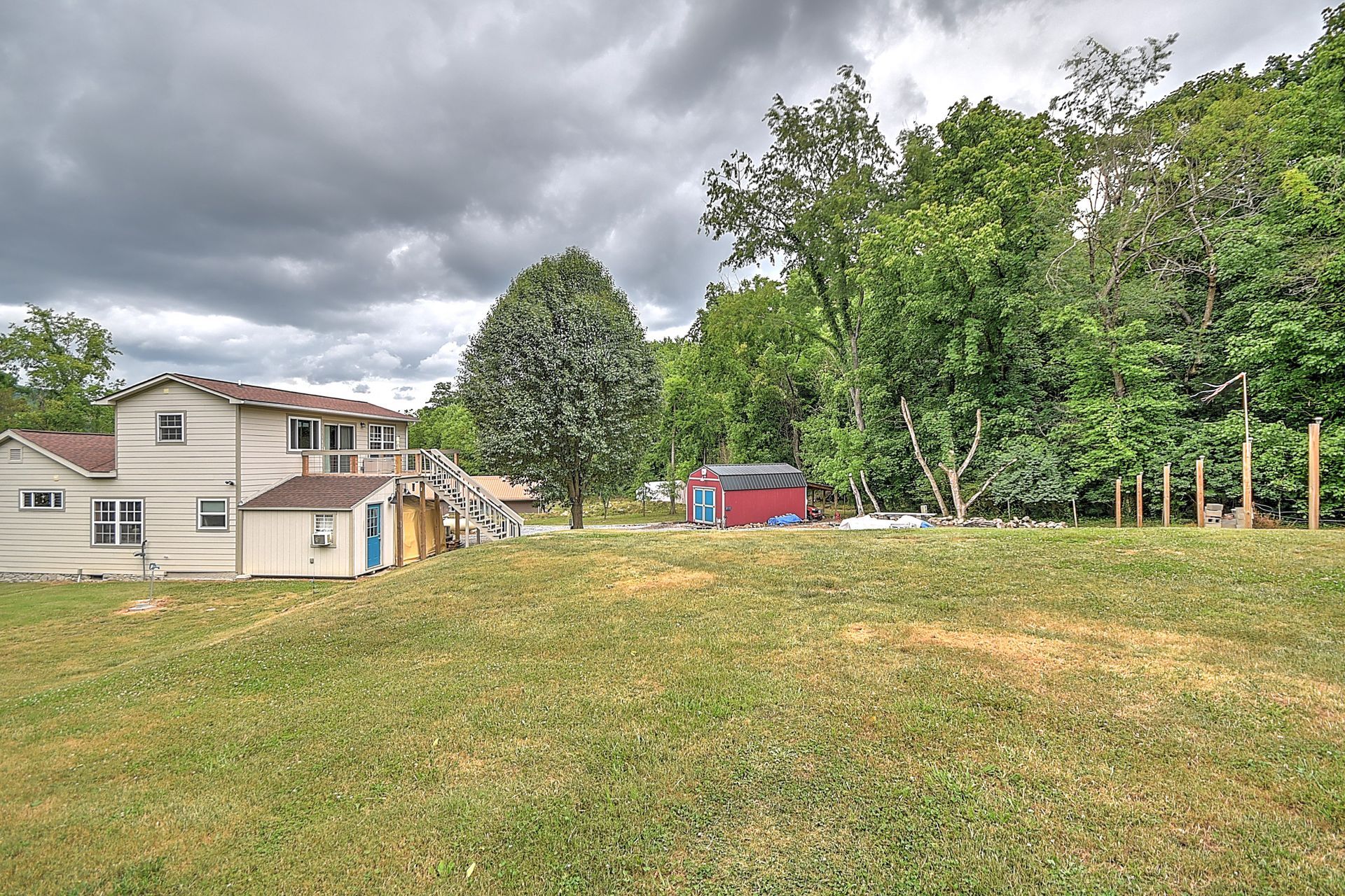 Grassy backyard beside a beige house, with a red shed and trees under a cloudy sky.