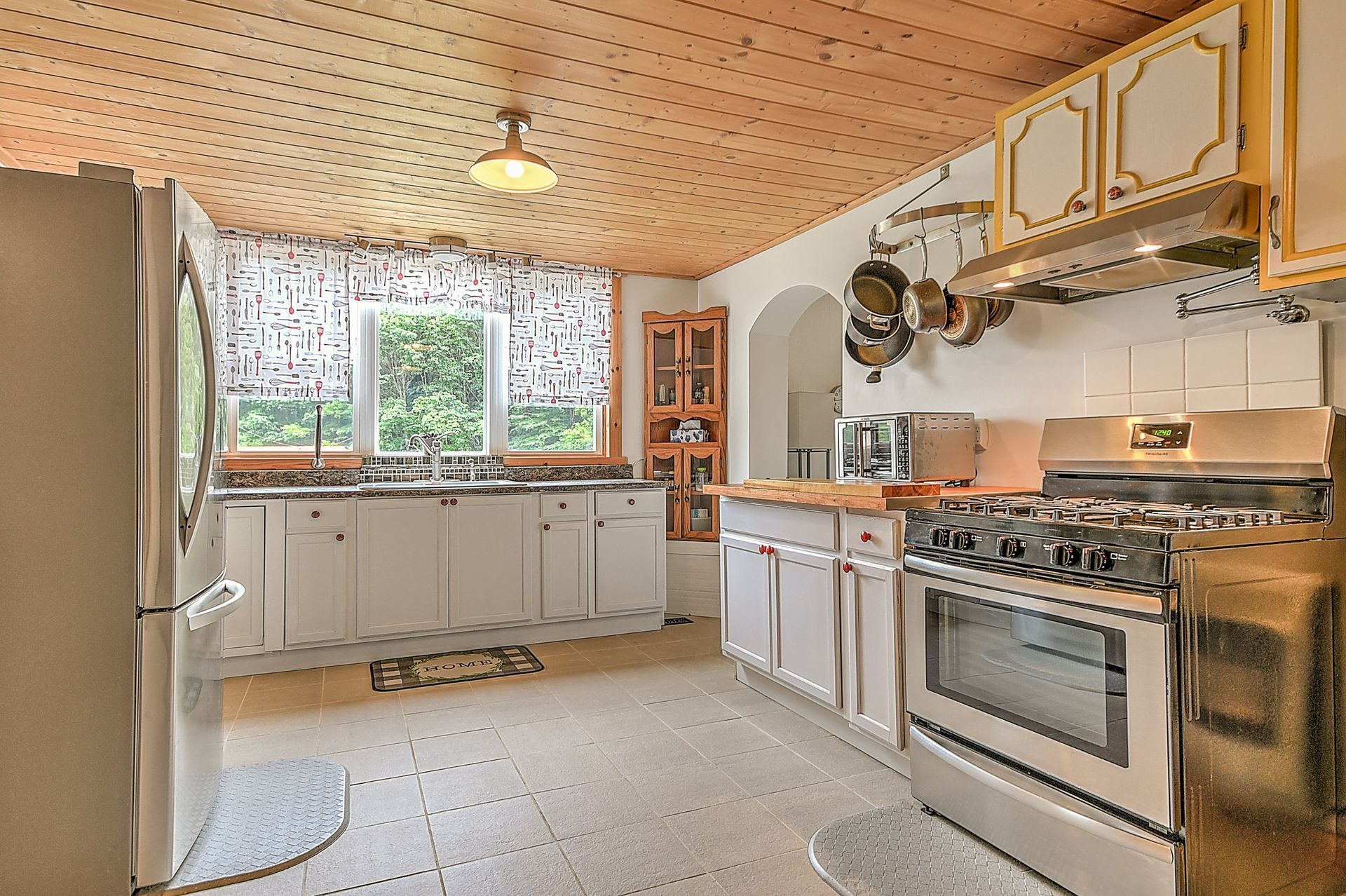 Bright kitchen with white cabinets, stainless stove, wood-paneled ceiling, and large window over the sink.