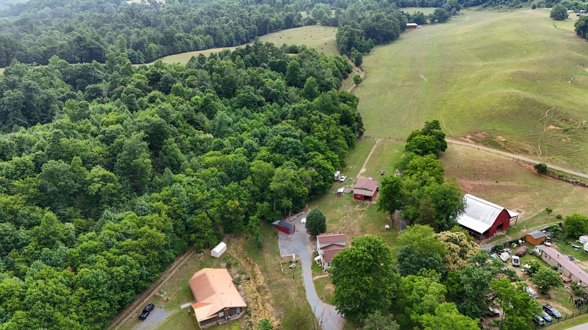 Aerial view of a rural farm with green forest, fields, and scattered houses along a winding dirt road.