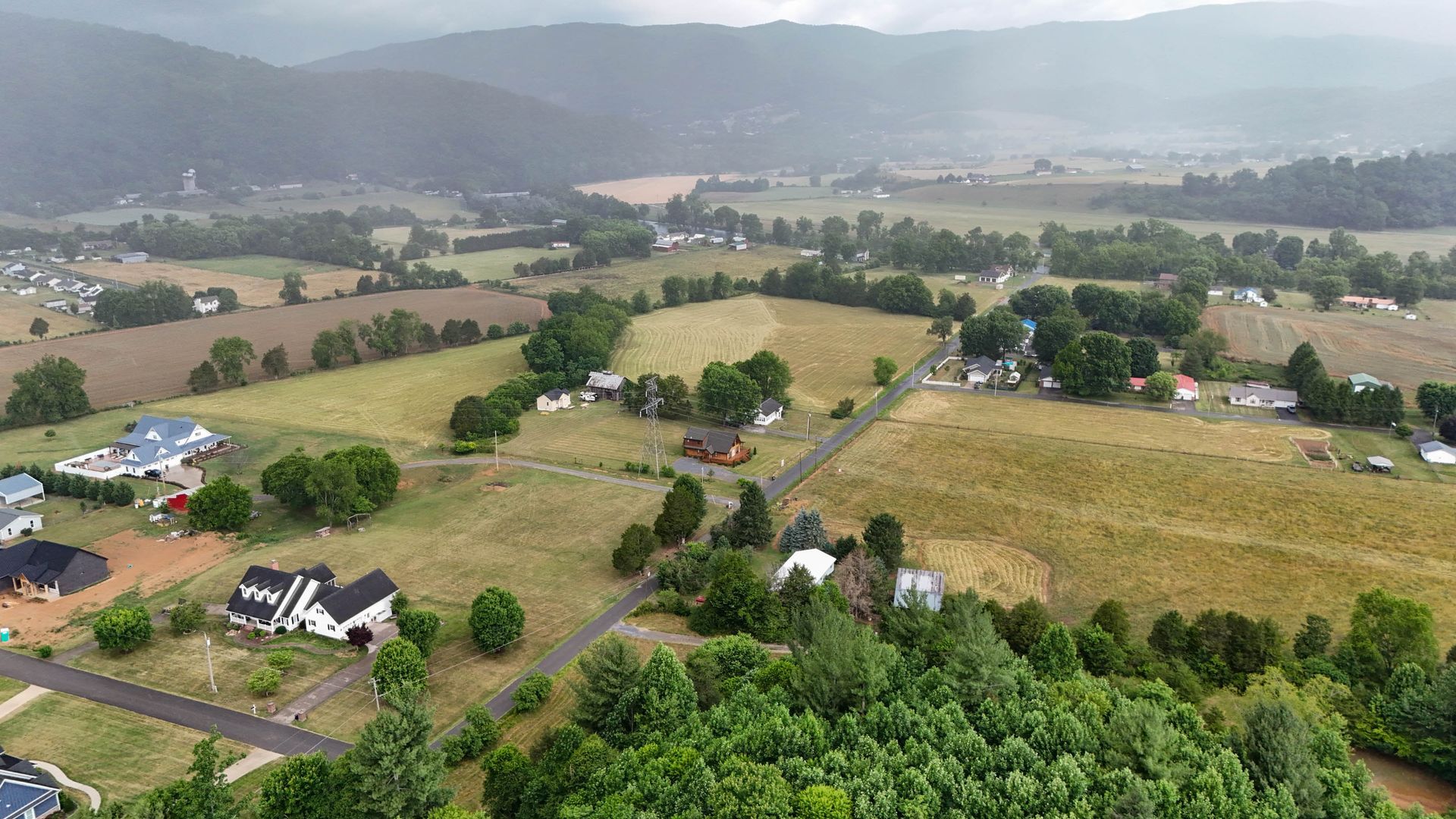 Aerial view of a rural valley with fields, scattered houses, trees, and misty hills in the distance