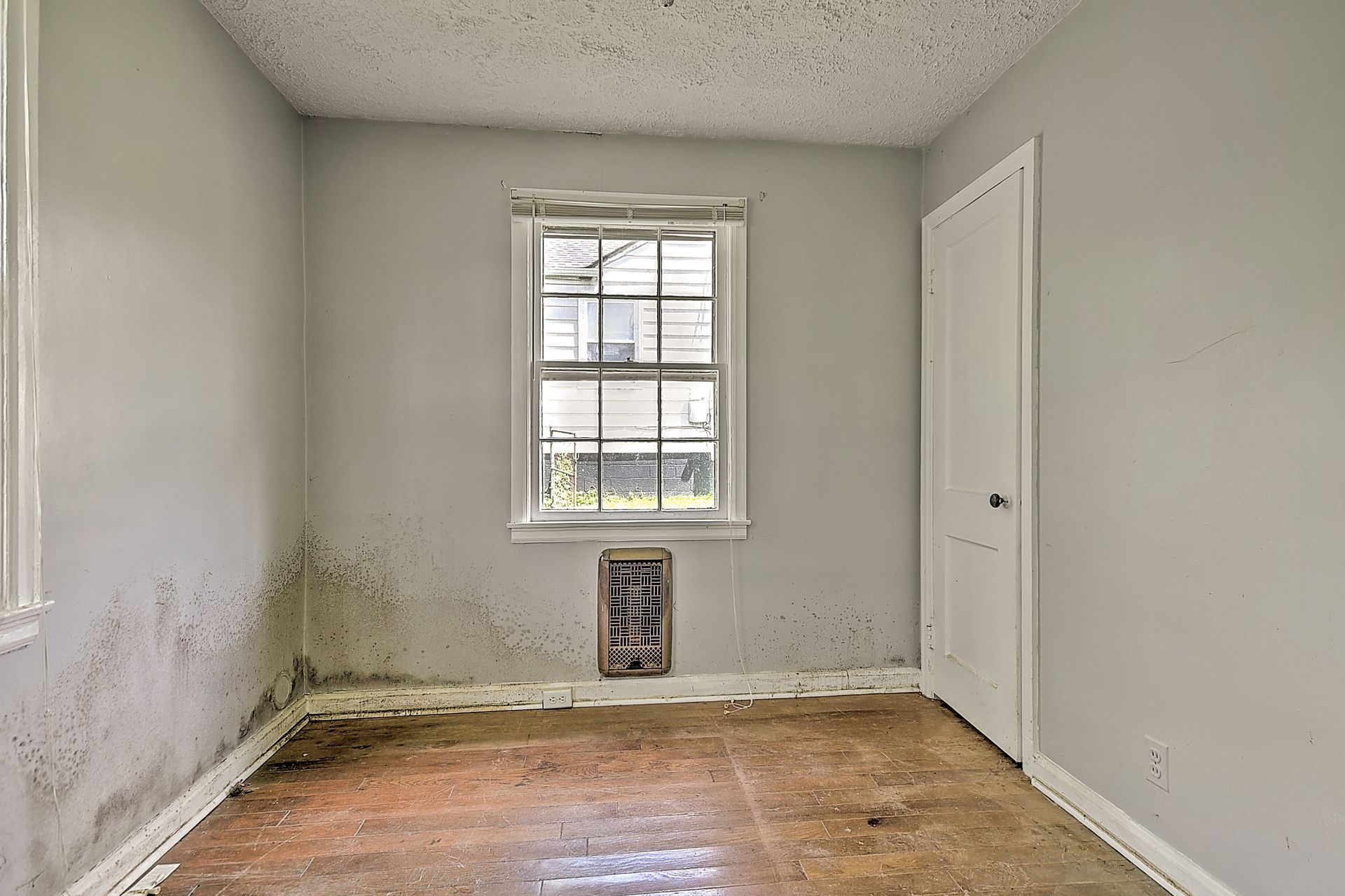 Empty small room with water-stained walls, bare floor, window, and closed door