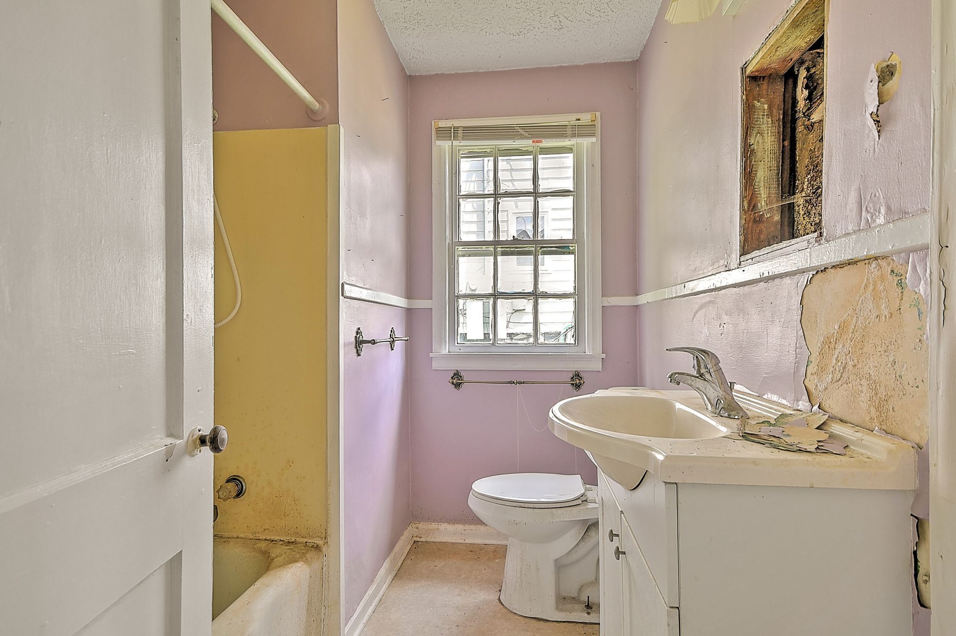 Small bathroom with torn pink walls, sink, toilet, and a window letting in daylight.