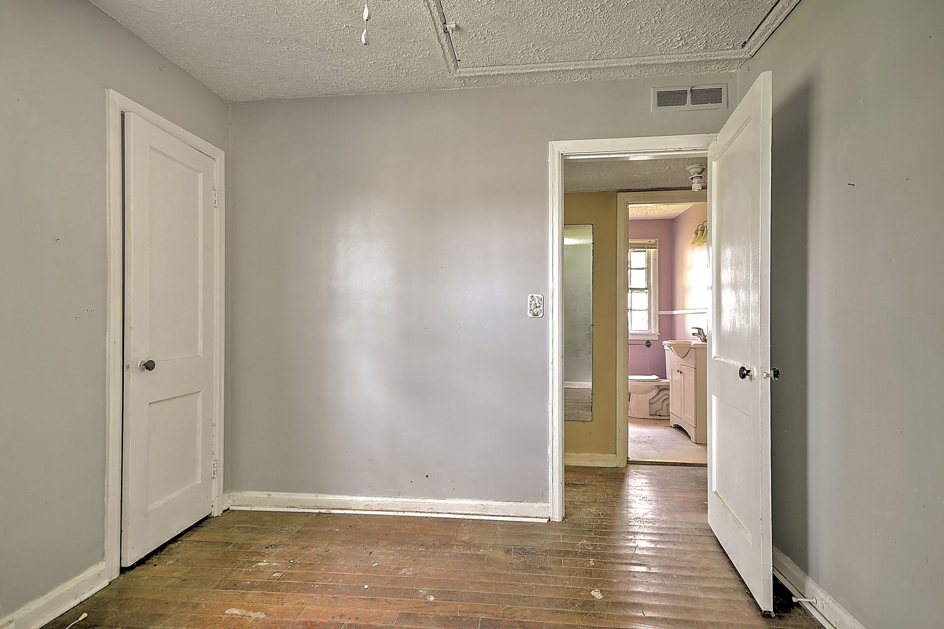 Empty gray room with worn wood floors and an open doorway to a brighter hallway