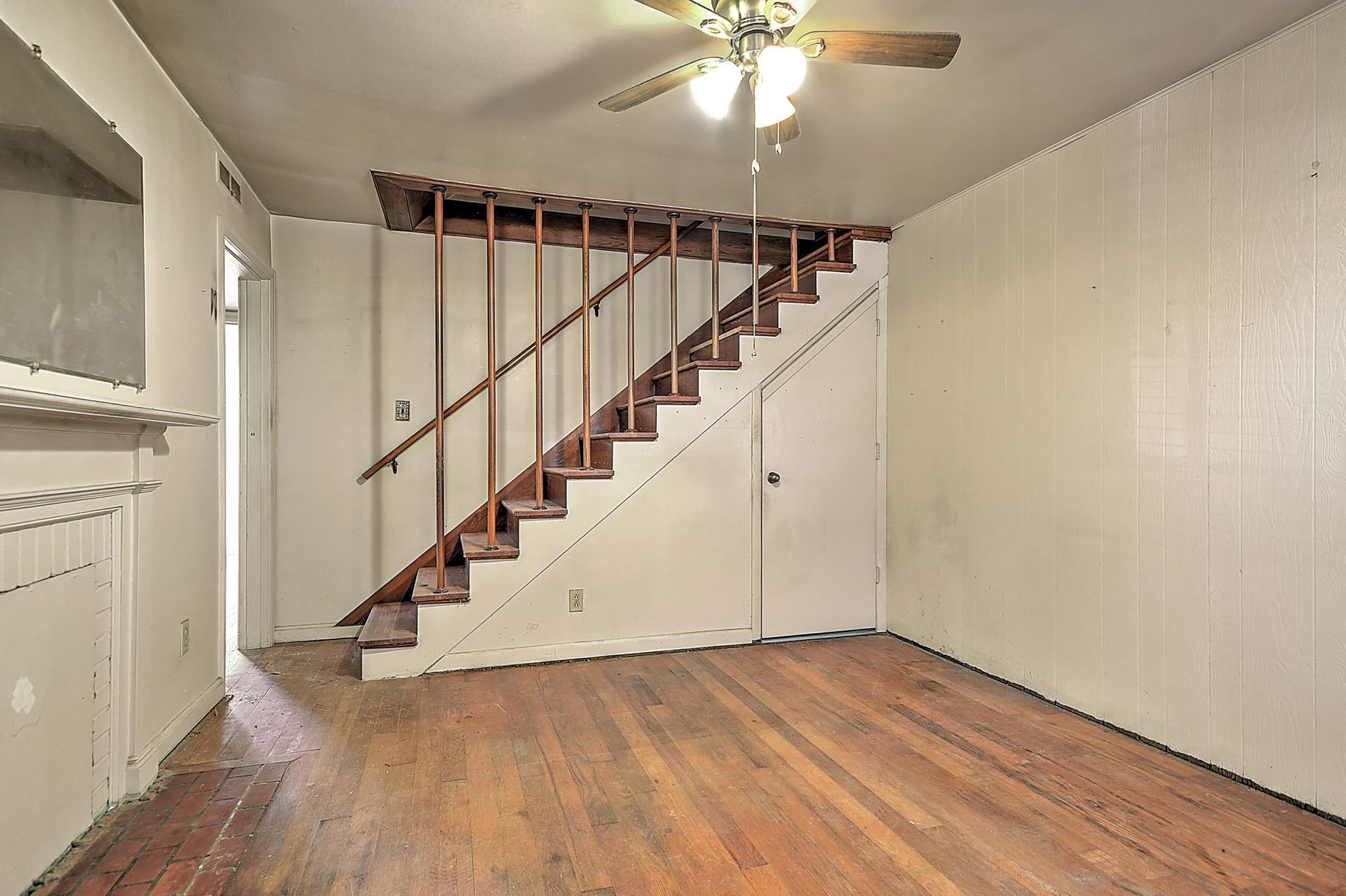 Empty room with wood floor, white paneled walls, and a staircase along the back wall under a ceiling fan.