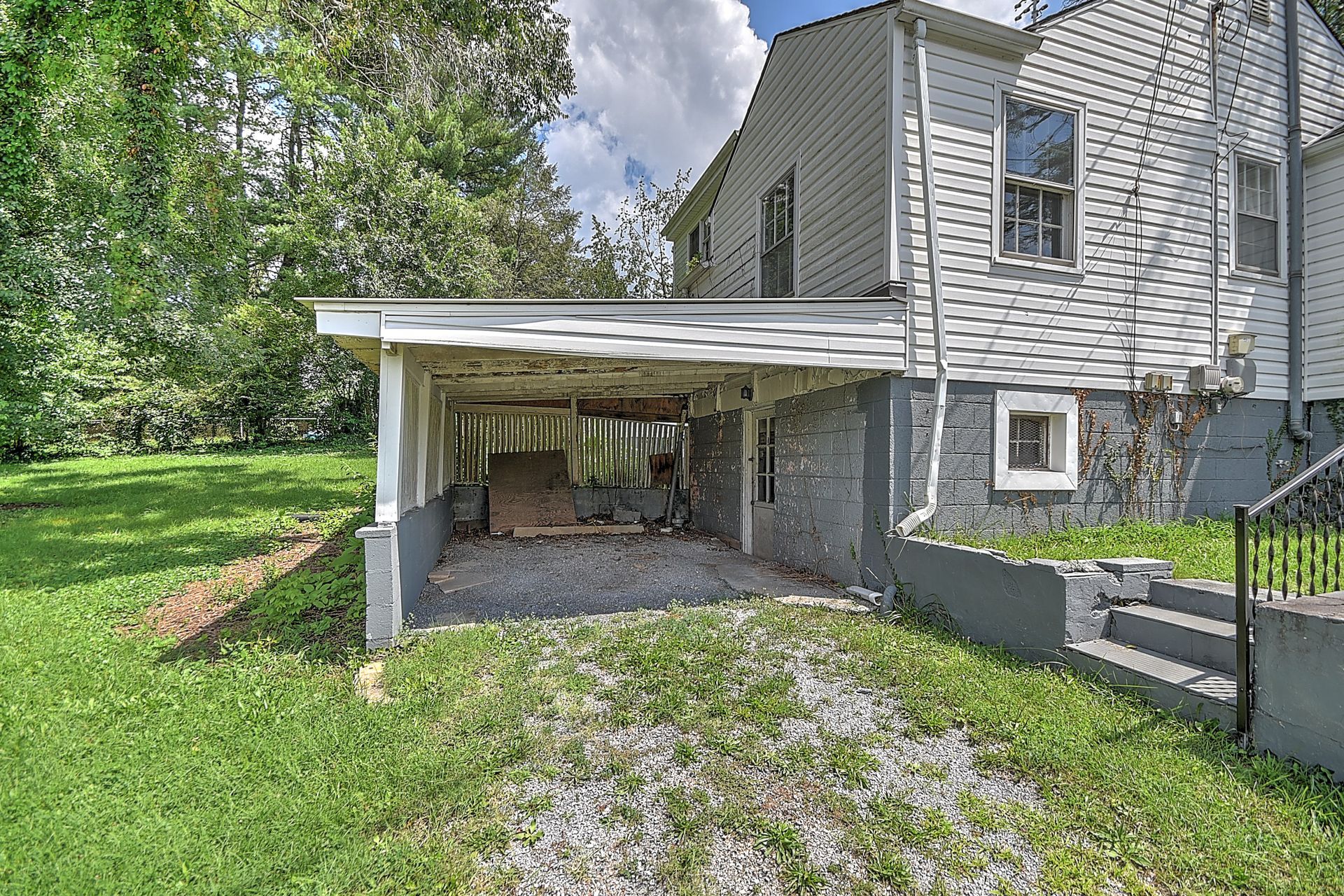 Covered carport beside a gray house with grassy yard and trees