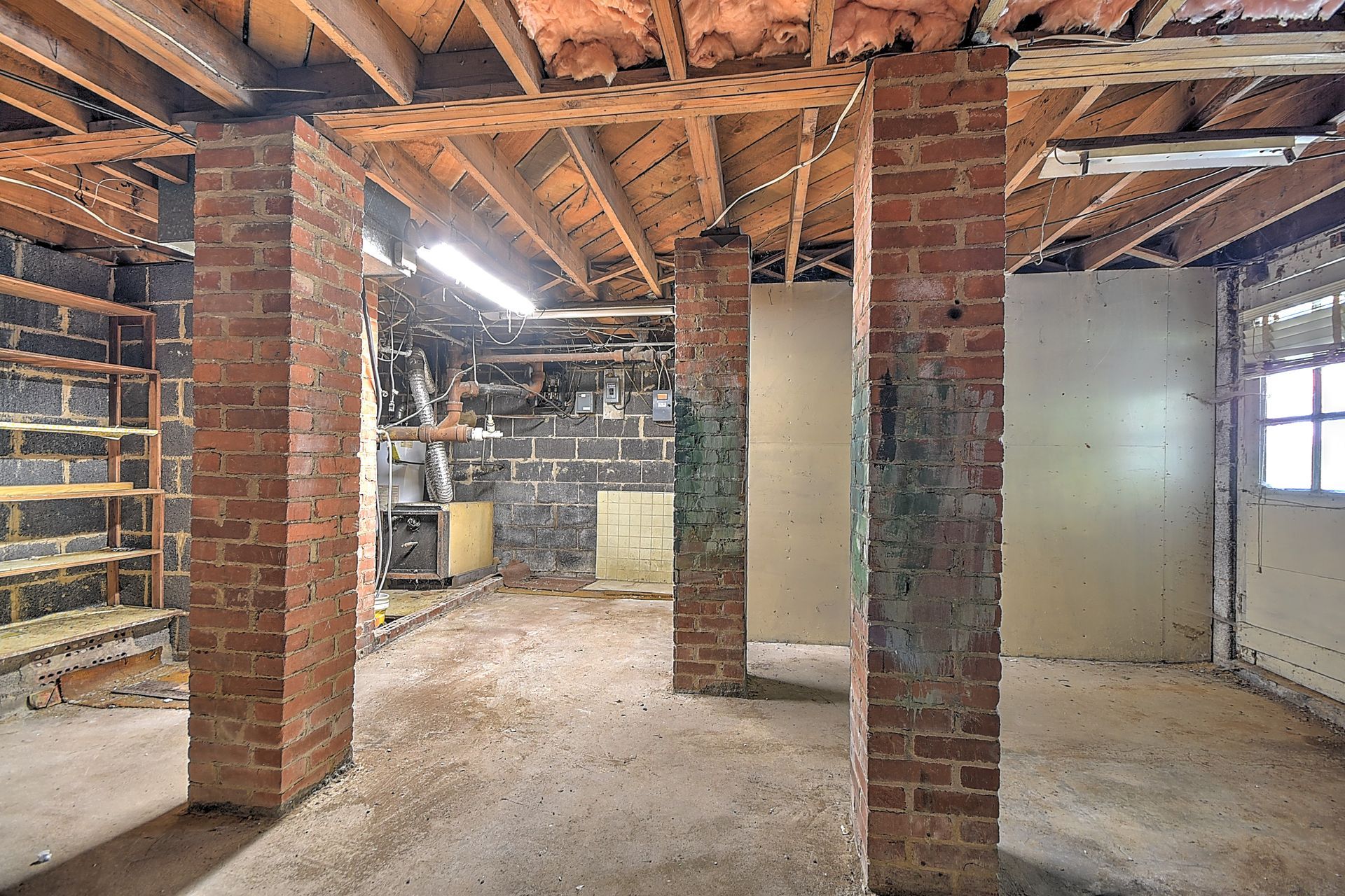 Unfinished basement with exposed wooden beams, brick pillars, and concrete floor under renovation.